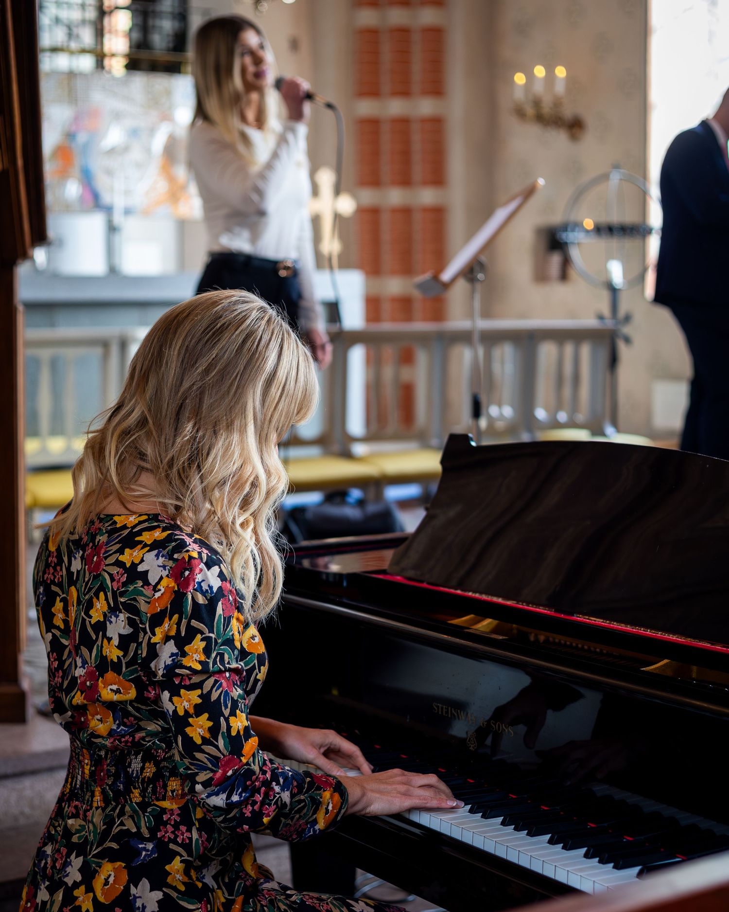 Musiker under dopceremoni i kyrka – pianist i blommig klänning och sångerska vid mikrofon.