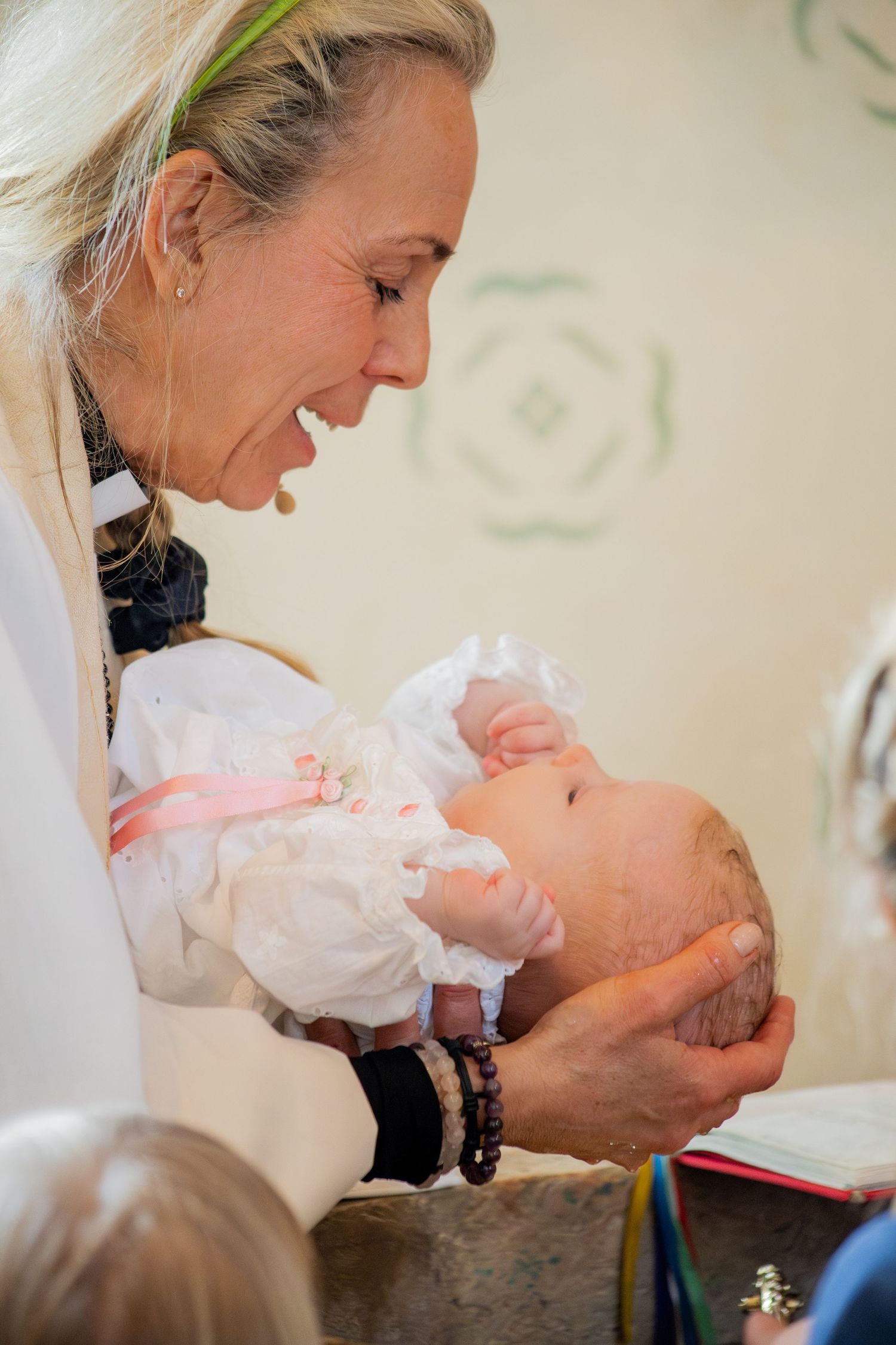 Präst håller nyfödd i dopklänning vid dopfunt, närbild under dop i kyrka.
