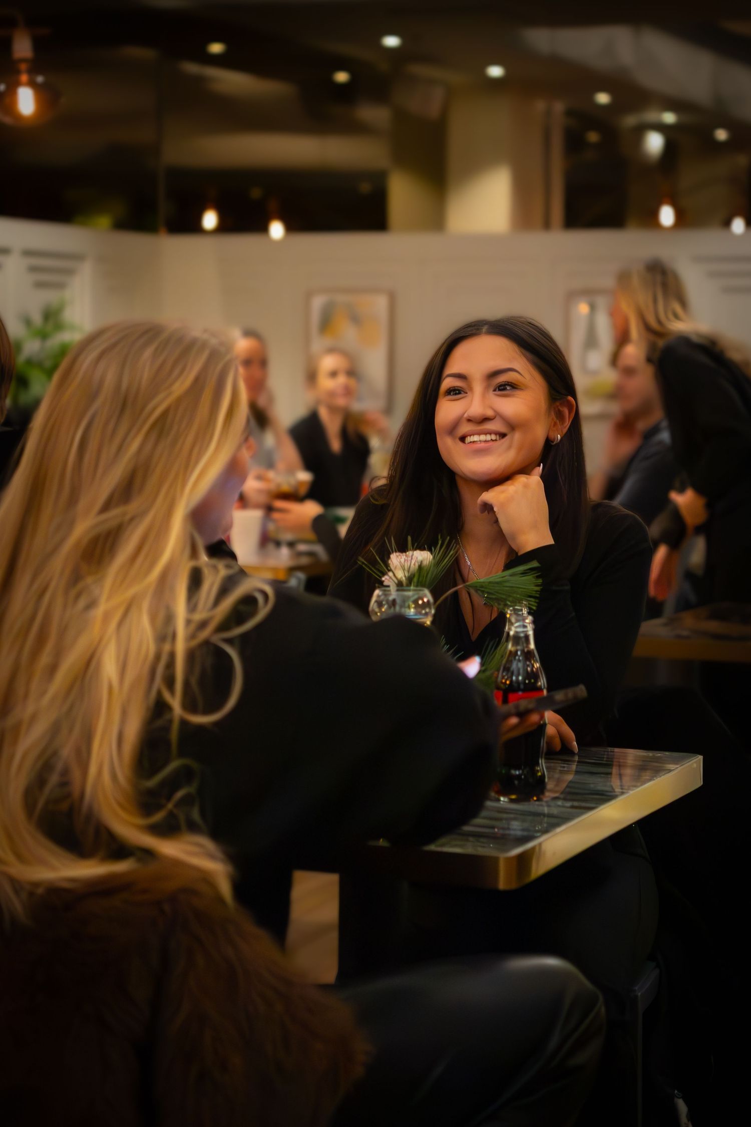 Två personer i livlig konversation vid restaurangbord med varm belysning och gäster i bakgrunden.