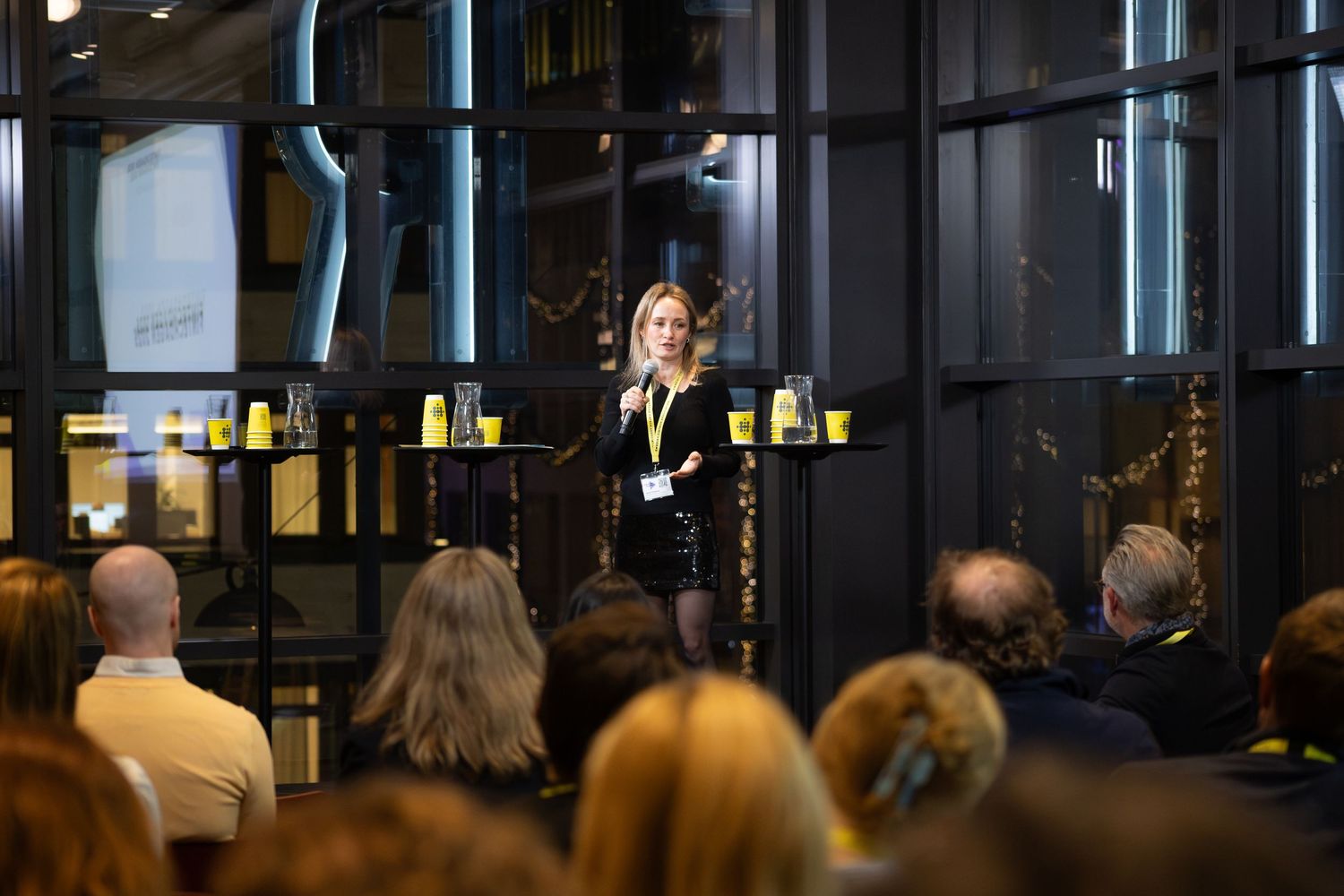 Talare på scen framför publik i glasad eventlokal, eventfotograf och företagsfotograf på konferens.
