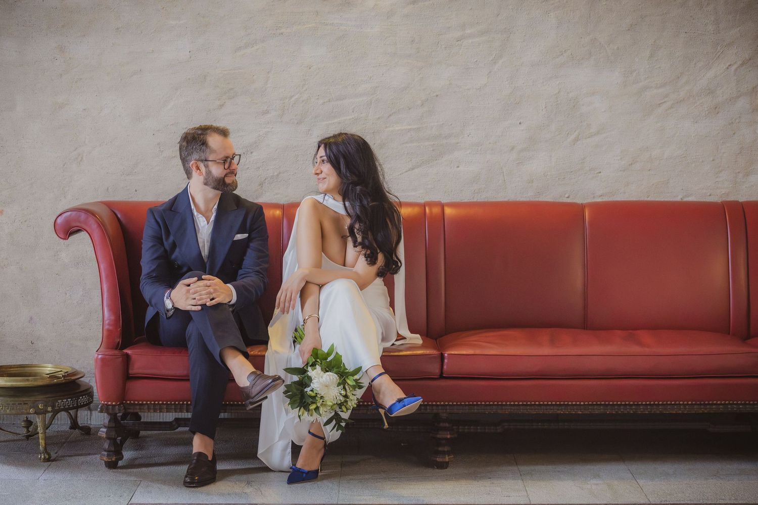Bride and groom sitting together on a red vintage sofa, smiling at each other, bride holding a white bouquet.