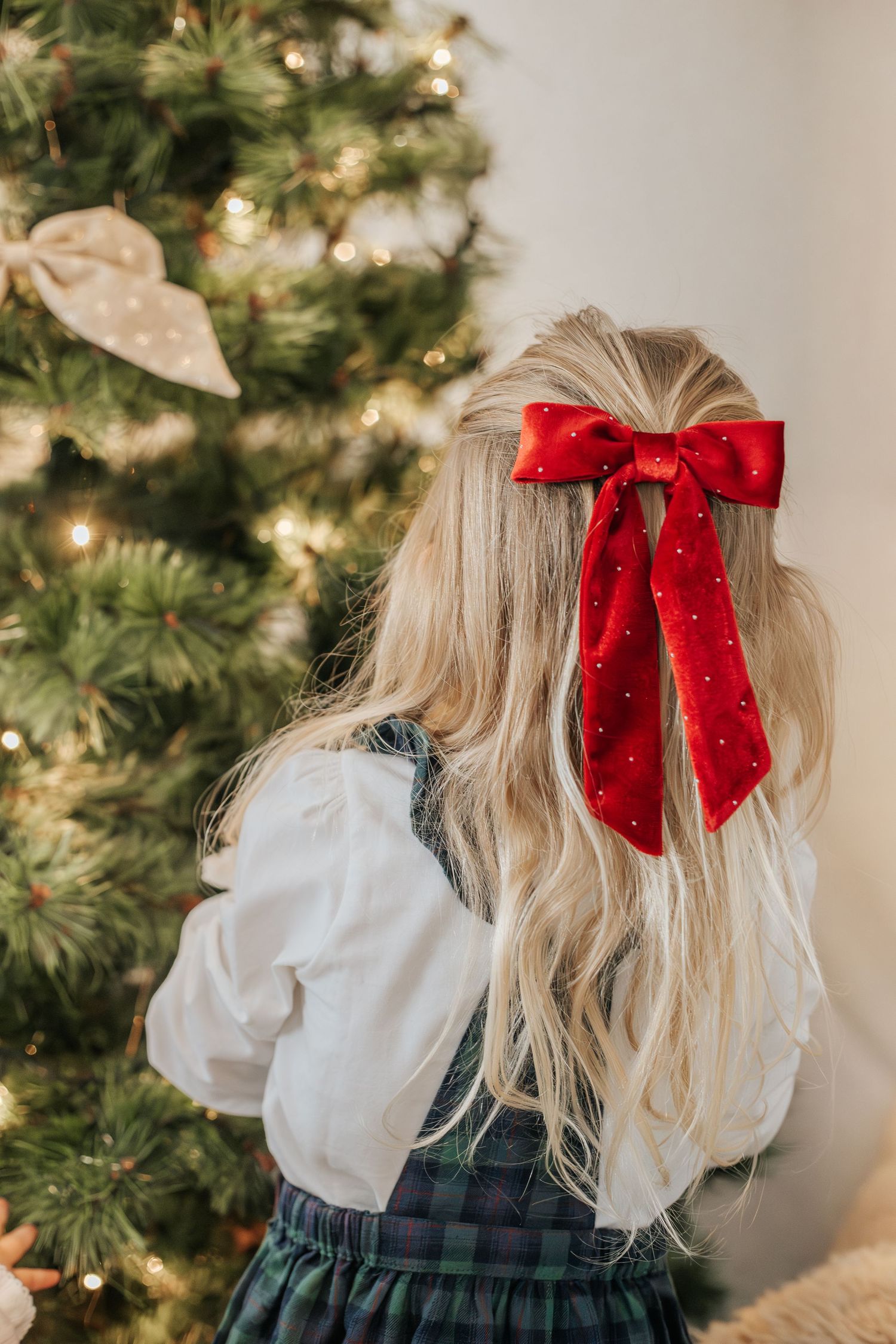 Barn med röd rosett i håret vid julgran, familjefotograf dokumenterar julporträtt inomhus.