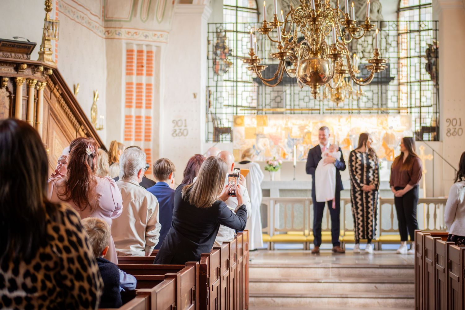 Dop i kyrka med familj och präst vid altaret, gäster i bänkar, dokumentär dopfotografering.