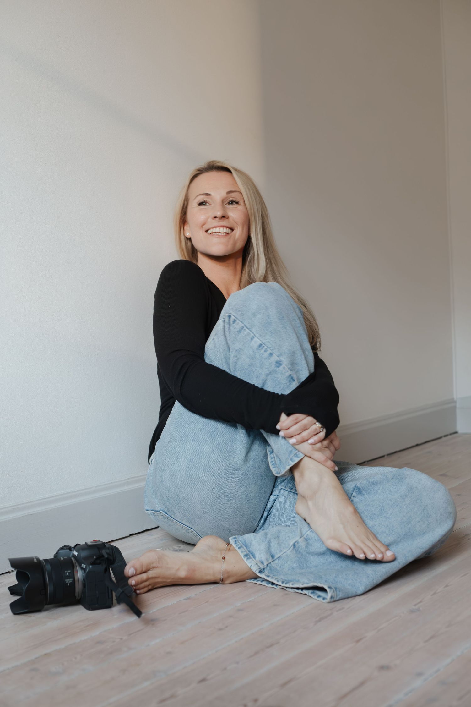 Fotograf i svart tröja och blå jeans sitter på golv bredvid kamera, fotografporträtt i naturligt ljus.