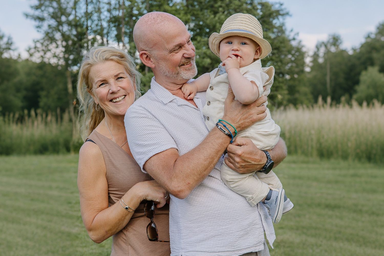 Äldre par med litet barn i famnen utomhus på äng, familjefotografering i naturligt ljus. Generationsfotografering.