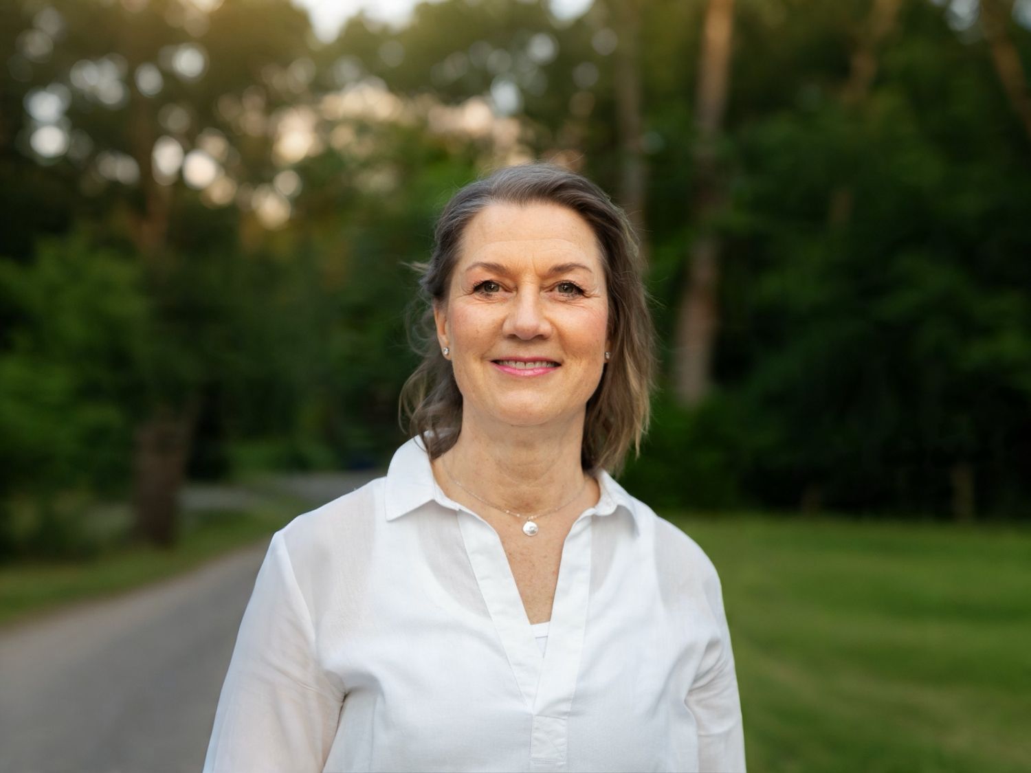 Smiling middle-aged woman with gray hair wearing a white shirt, standing outdoors with green trees in background.