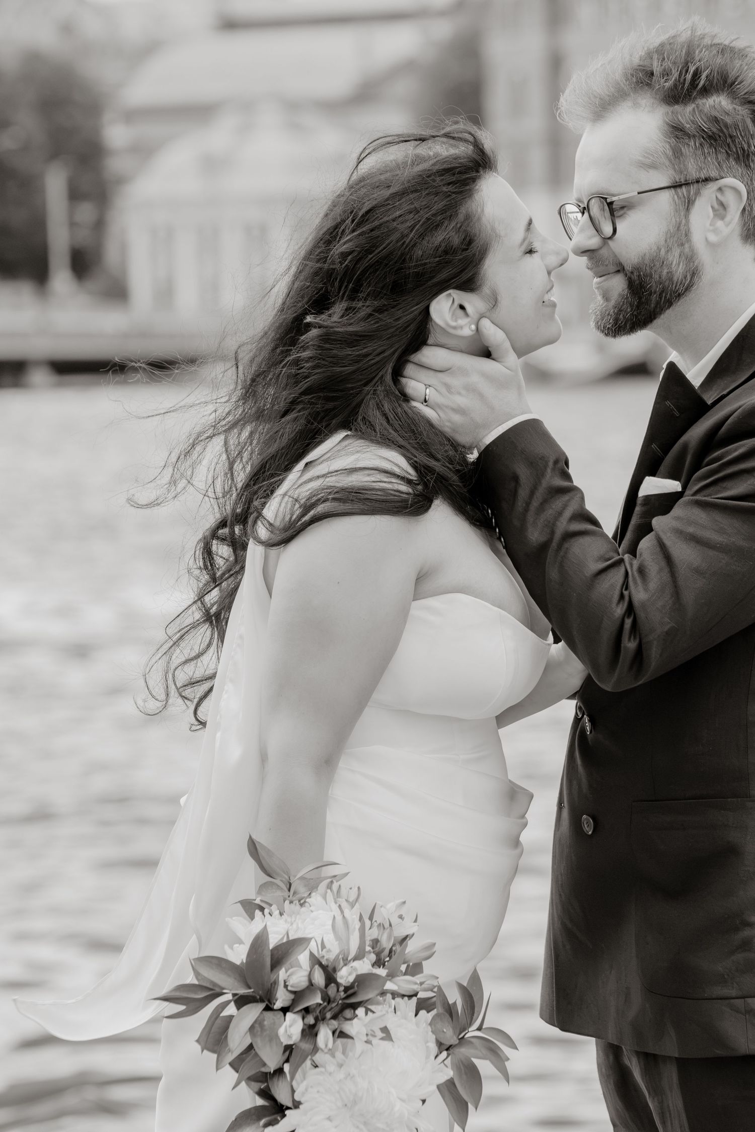 Black and white photo of a bride and groom sharing a romantic moment by the water, with her bouquet in hand.