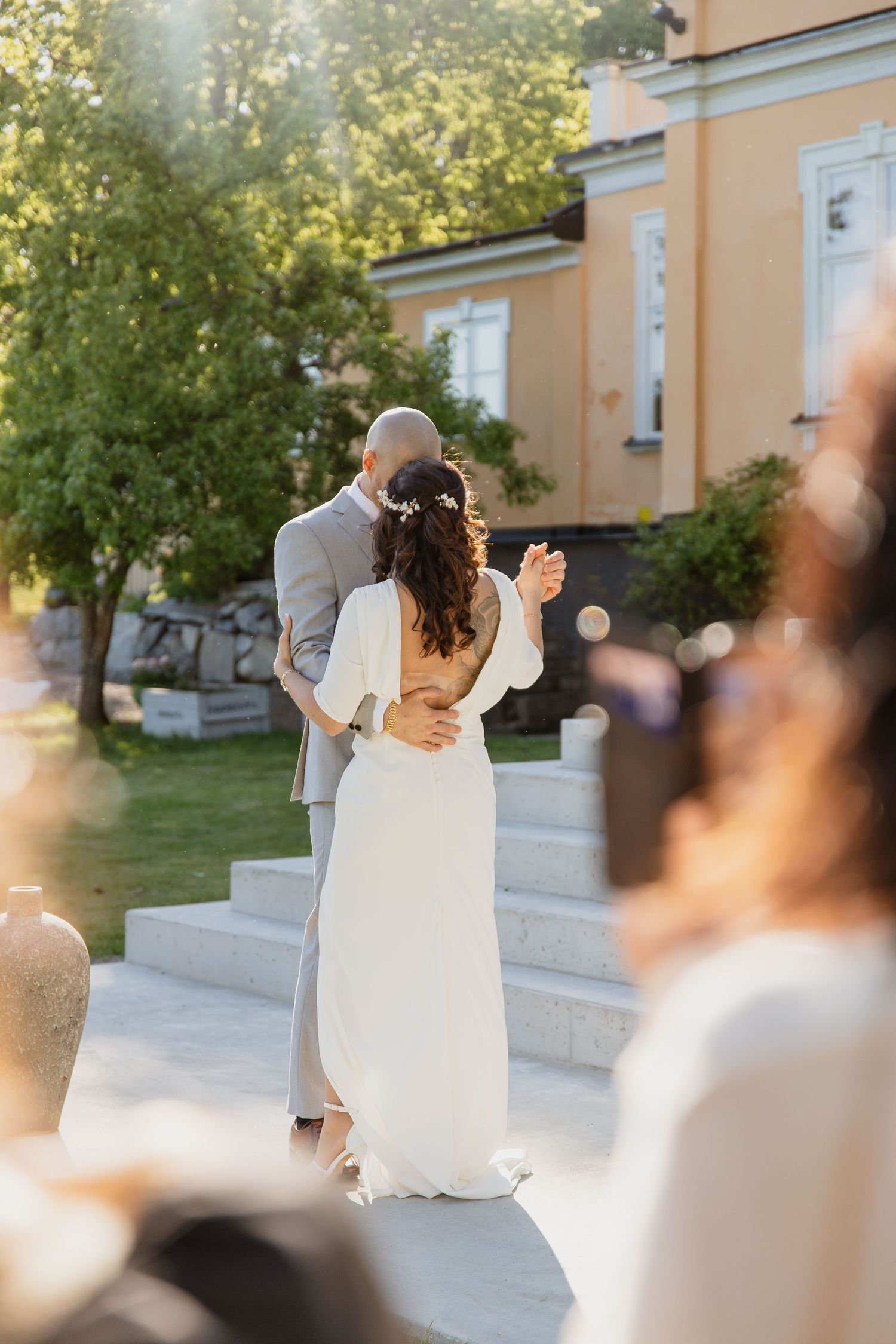 Brudparomfamnar varandra utomhus vid kyrktrappa efter dop, kvällsljus och gäster i bakgrunden, porträtt efter bröllopsceremoni. Bröllopsvals