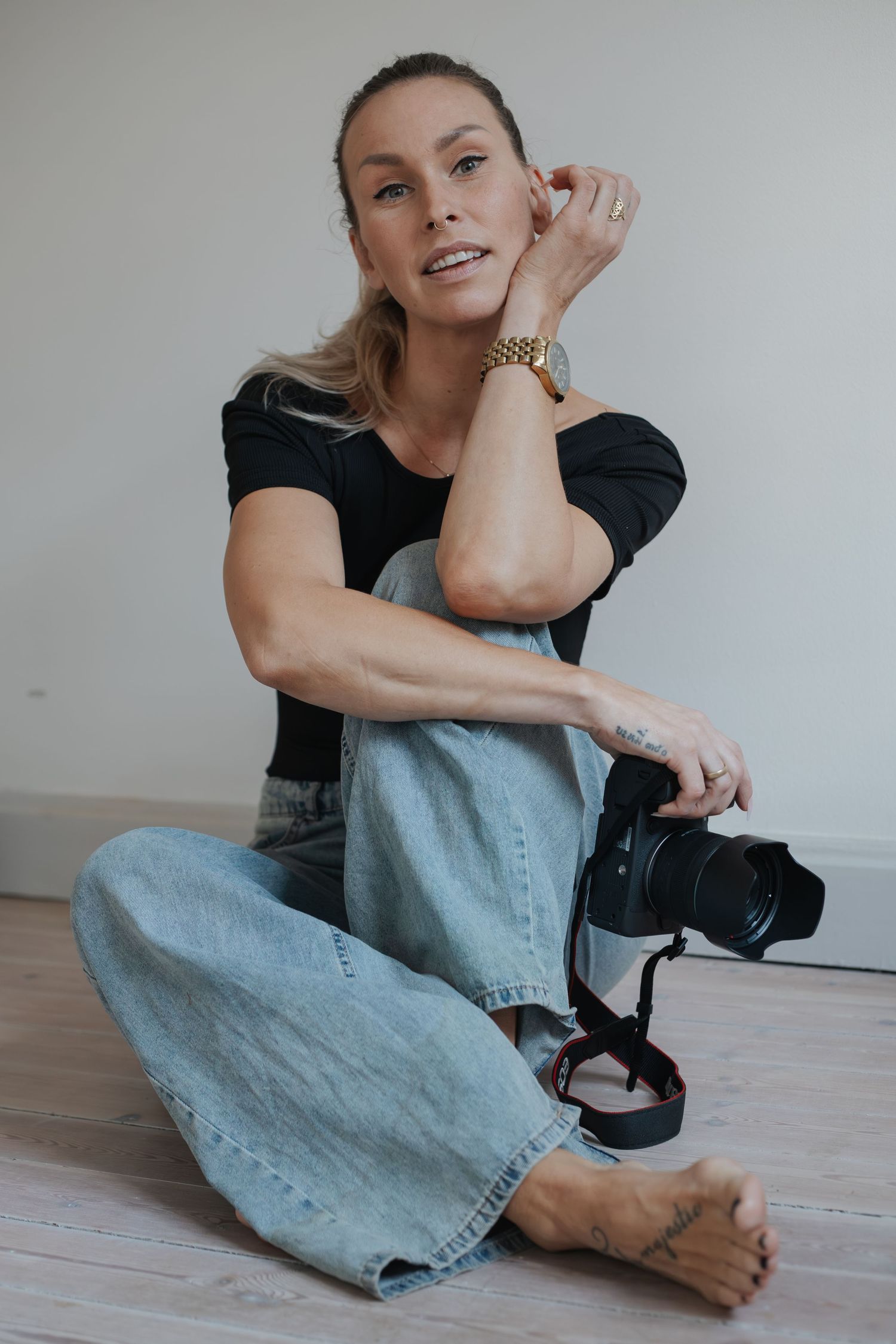 Fotograf i svart topp och blå jeans sitter på golvet med kamera, porträtt av företagsfotograf i studio.