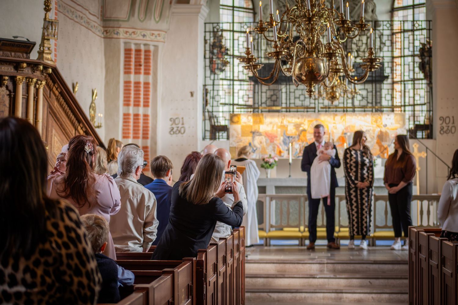 Dopceremoni i kyrka med präst och familj vid altaret, gäster sitter i bänkar, dopfotografering i kyrkomiljö.