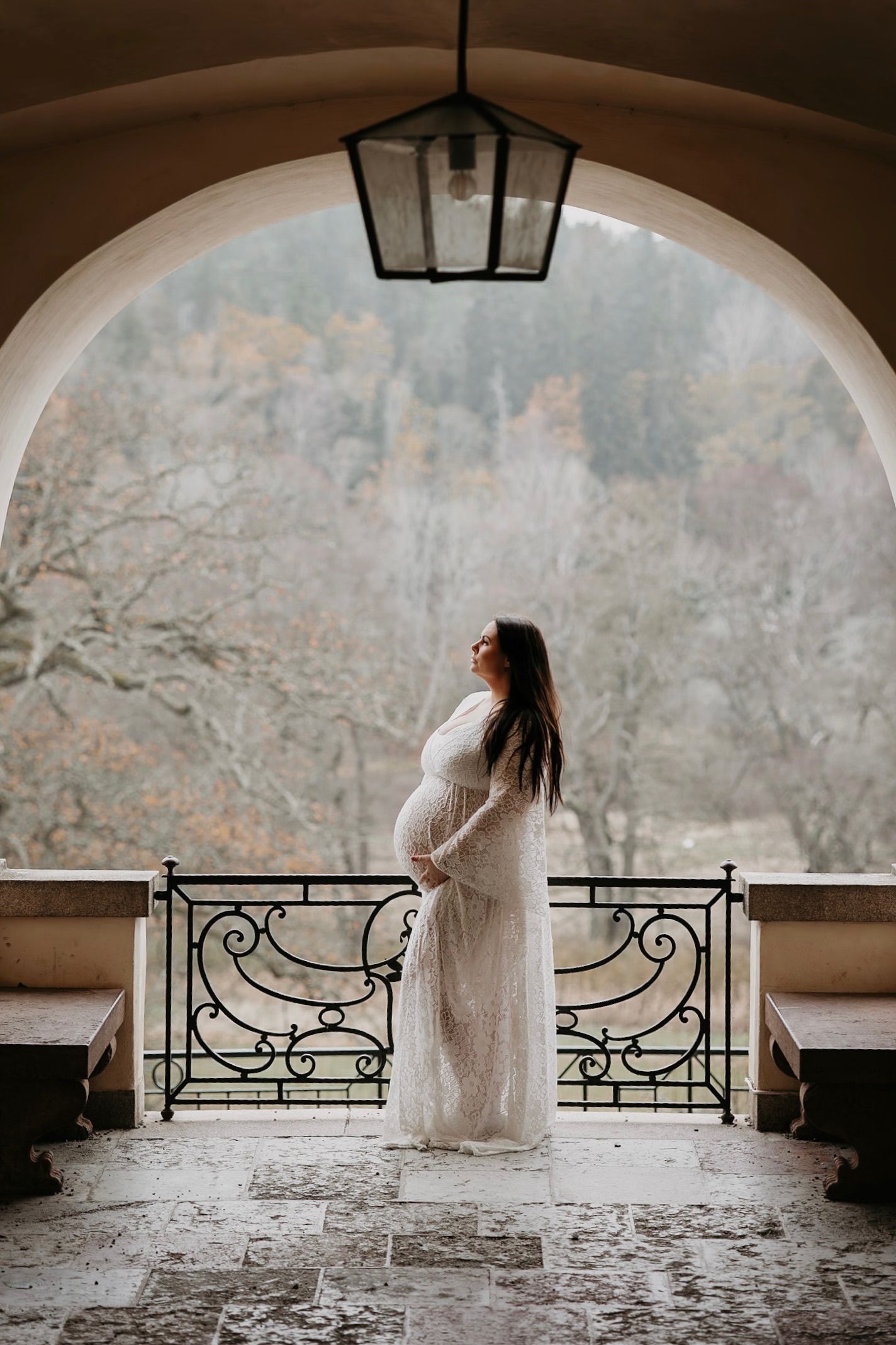 Gravid kvinna i vit klänning står under en stenvalv med vinterlandskap i bakgrunden – fotografering i graviditetens slutskede.