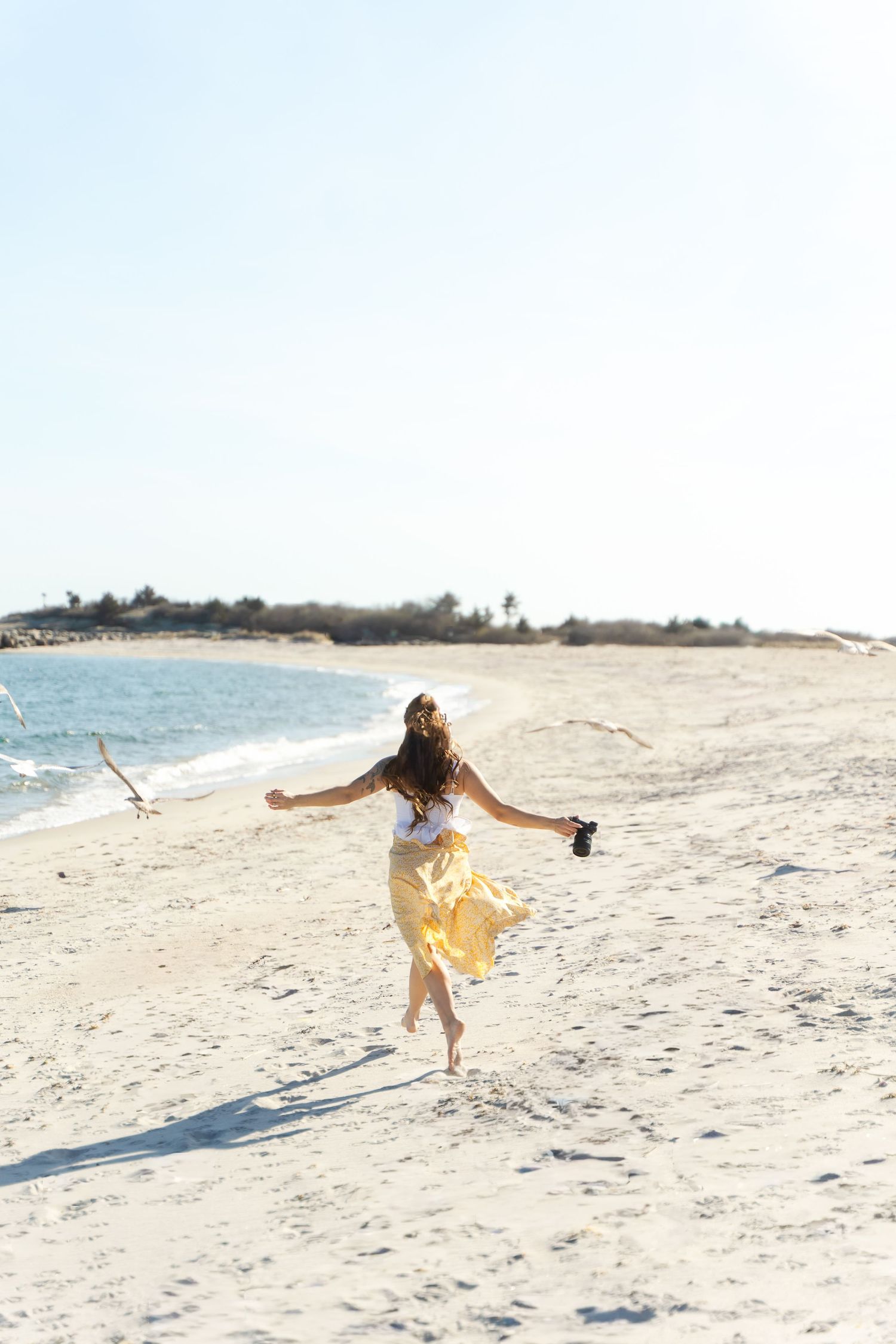 Woman in yellow skirt running freely on a sunny sandy beach near the ocean, holding shoes with arms outstretched. Ocean Beach Connecticut