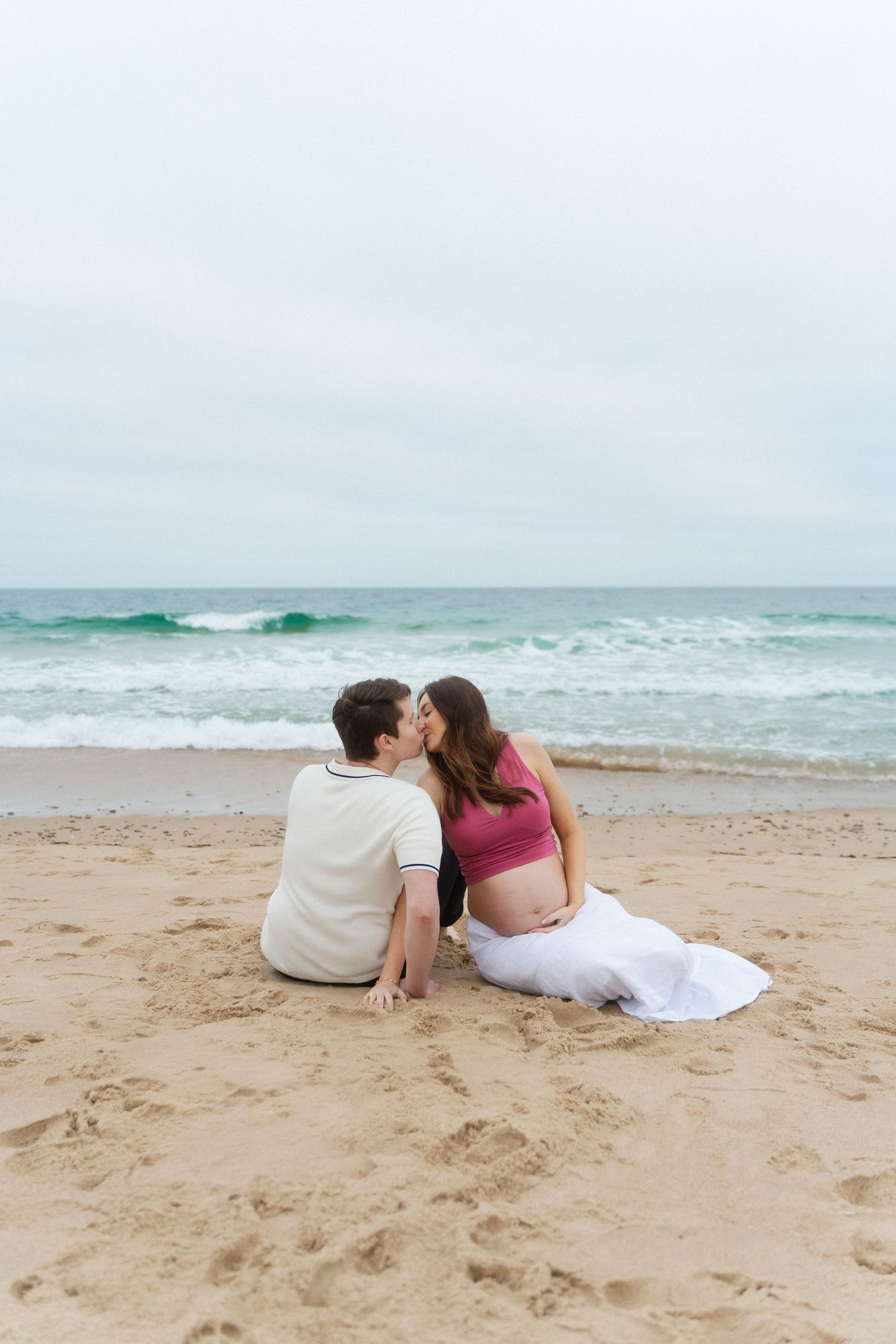 Couple sitting on sandy beach kissing, pregnant woman in pink crop top with white skirt, ocean waves in background.