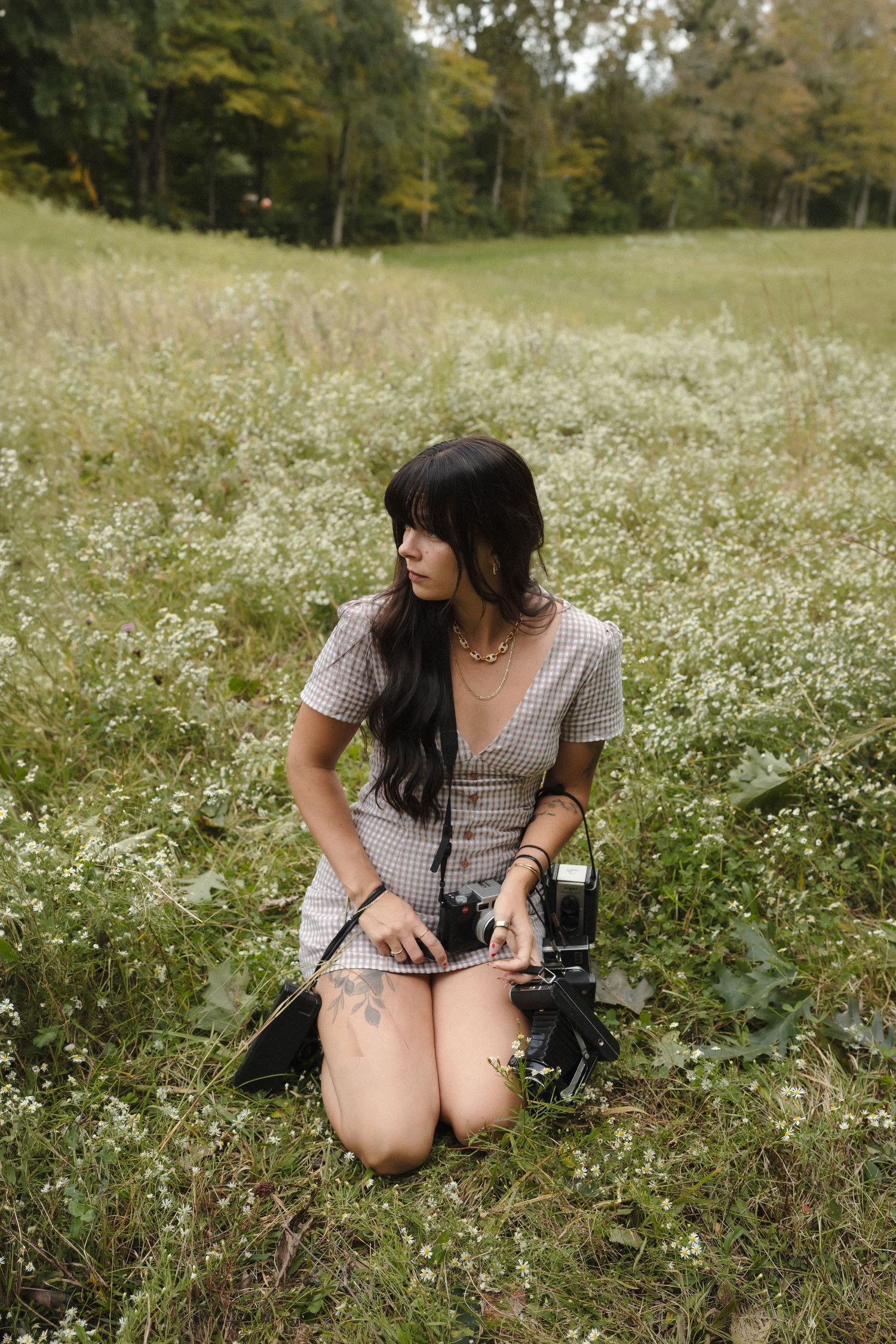 Woman kneeling in a wildflower field holding a vintage camera, wearing a plaid mini dress, with forest in the background.