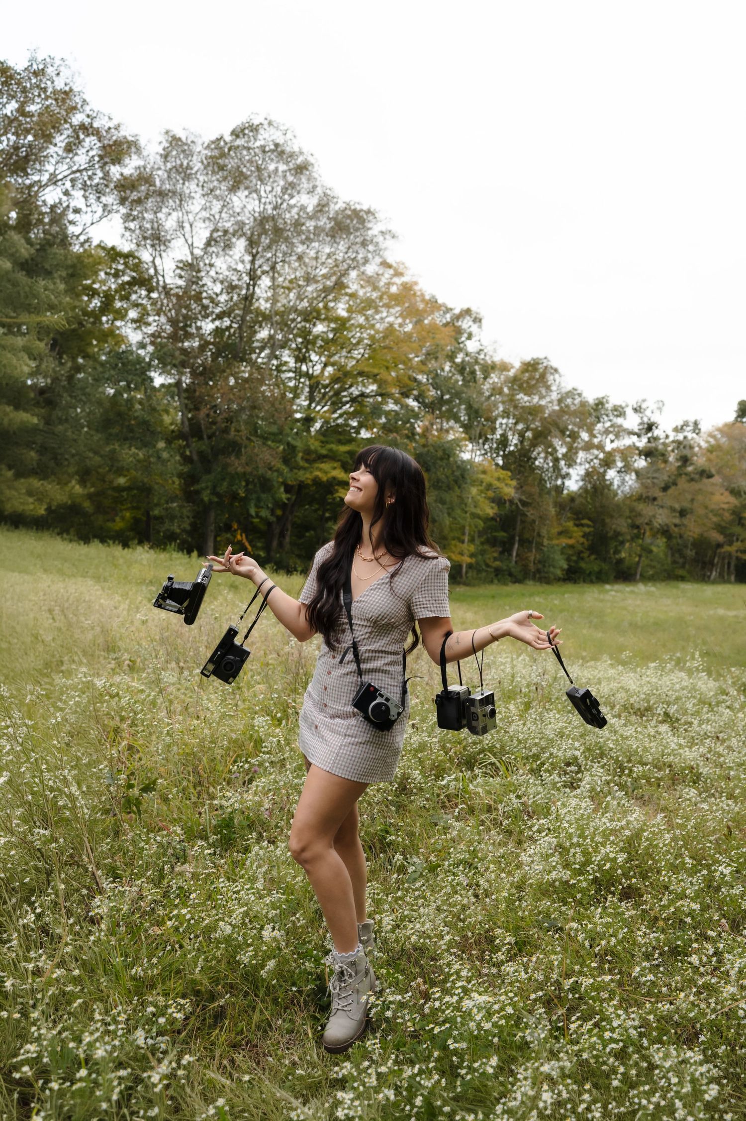 Woman holding multiple cameras in a wildflower field, wearing a gray dress and boots, surrounded by lush green trees.
