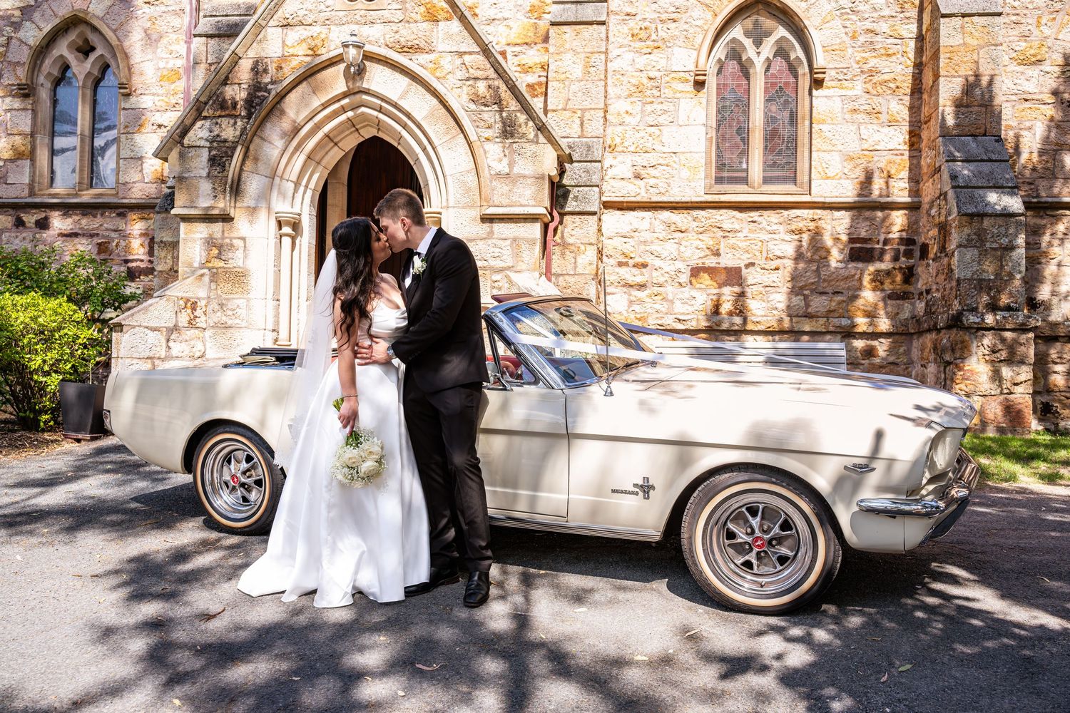 Newlyweds pose for romantic photos outside historic stone church with vintage car.