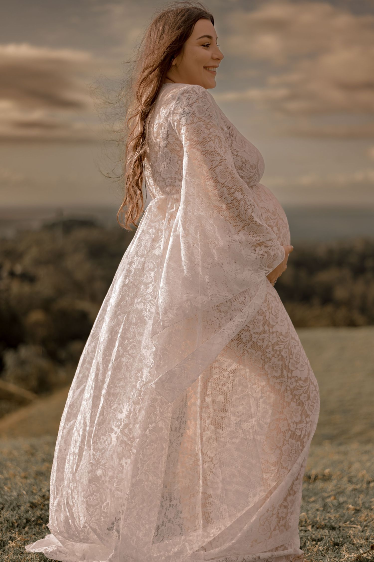Ethereal maternity photo of woman in lace dress against dramatic sunset sky.