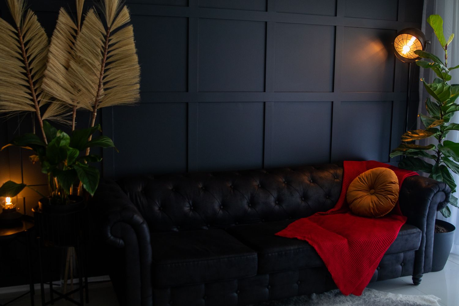 A cozy dark interior space with potted plants, warm lighting, and red accent pillows on gray furniture.