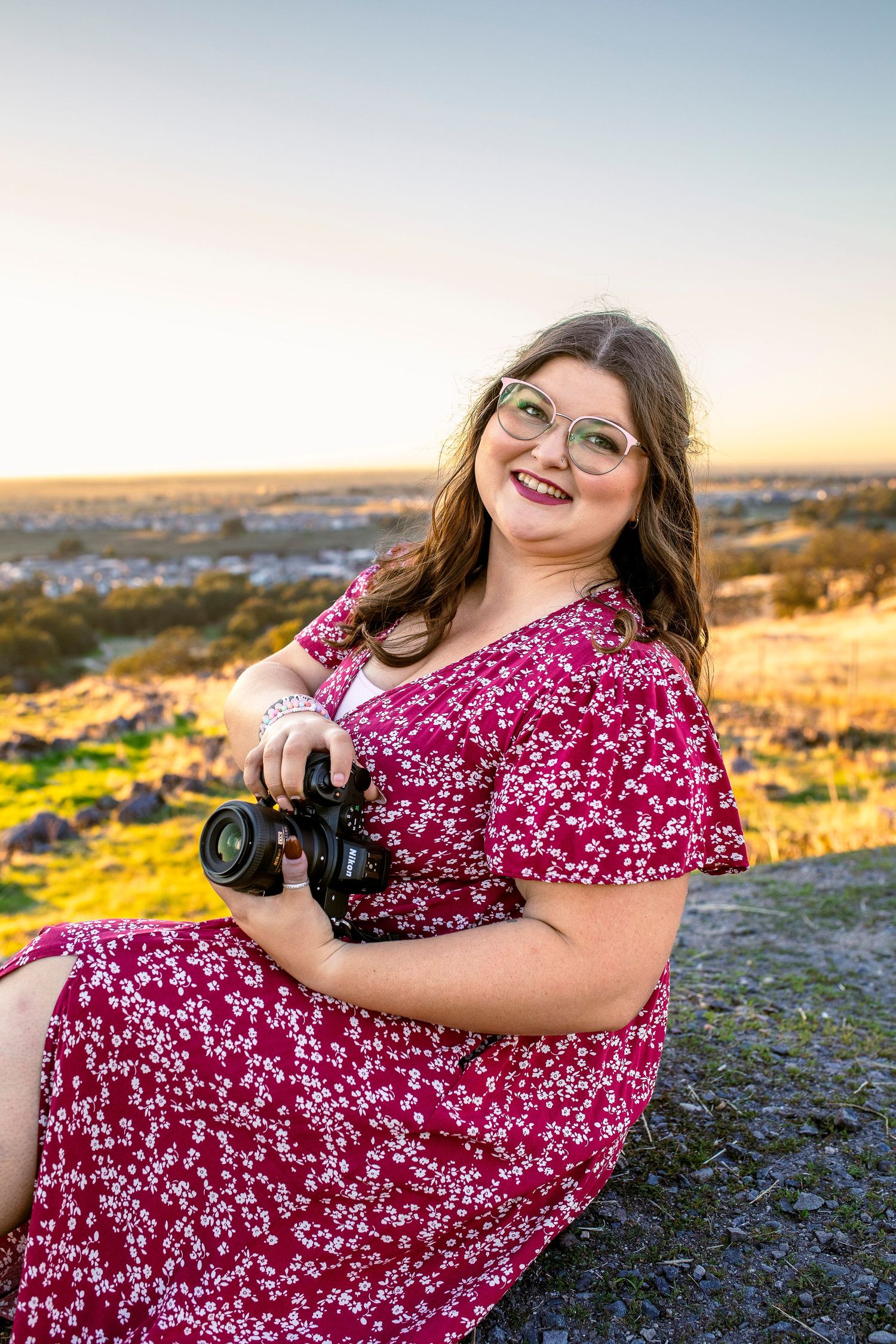Photographer in floral red dress poses with camera during golden hour sunset on rocky terrain.