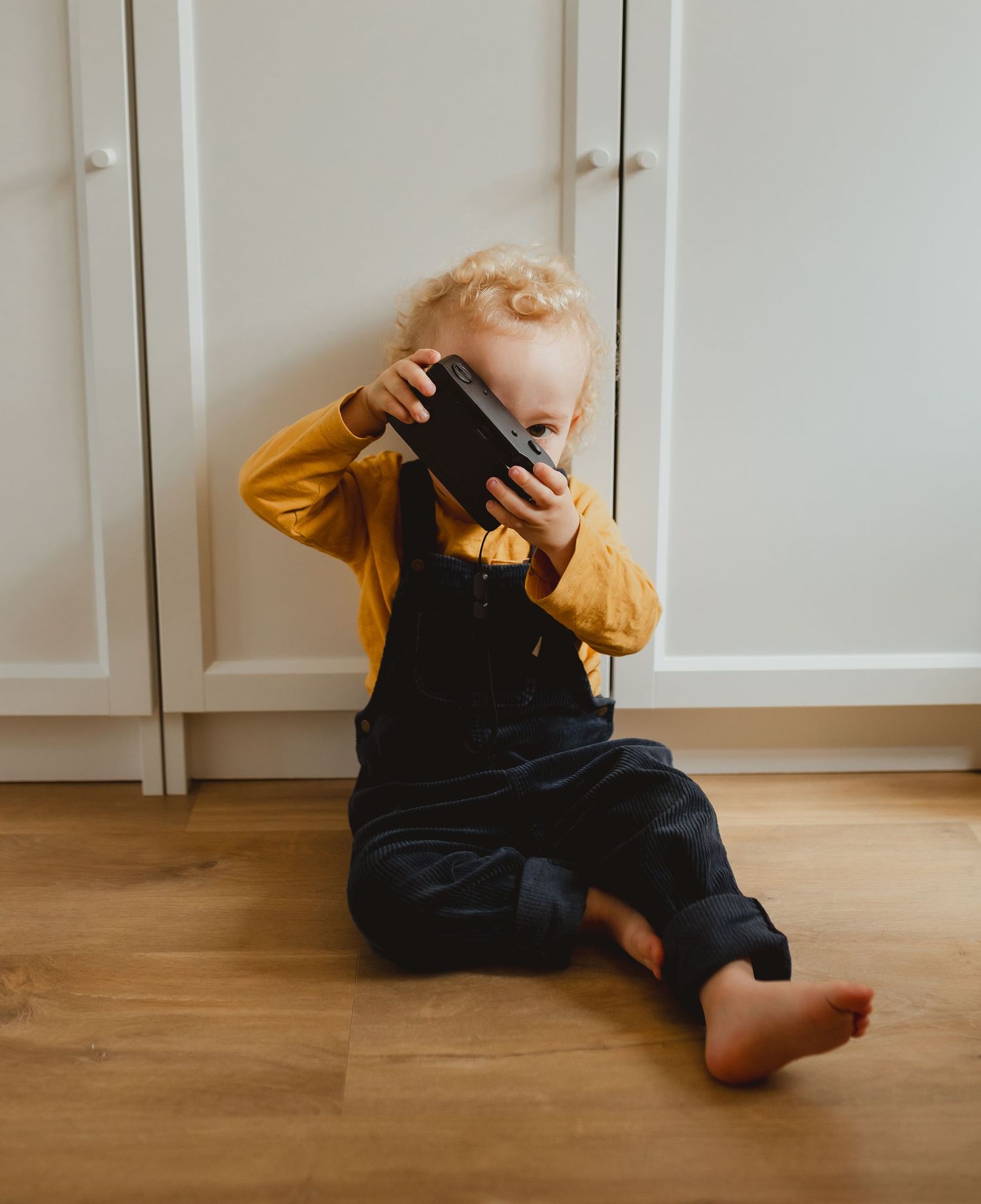 Small child in dark overalls and mustard yellow sweater sitting on wooden floor covering face with hands.