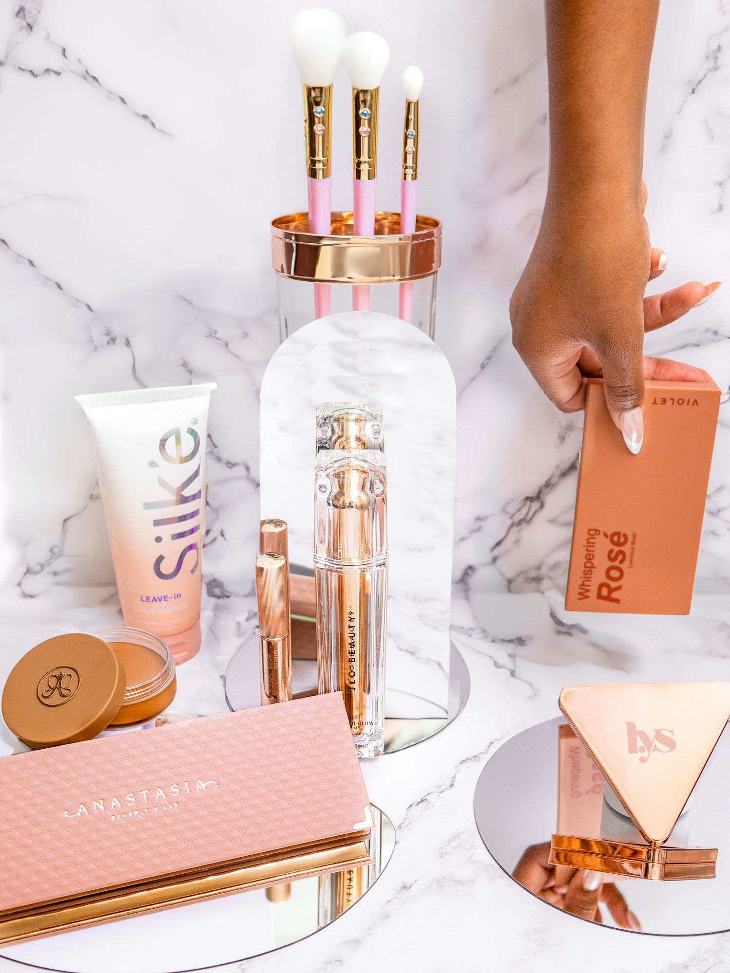 Luxury beauty products in rose gold tones arranged on white marble with makeup brushes.