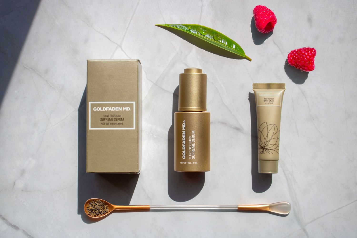 Beauty products with gold packaging arranged on marble surface with rose petals and green leaf accents.