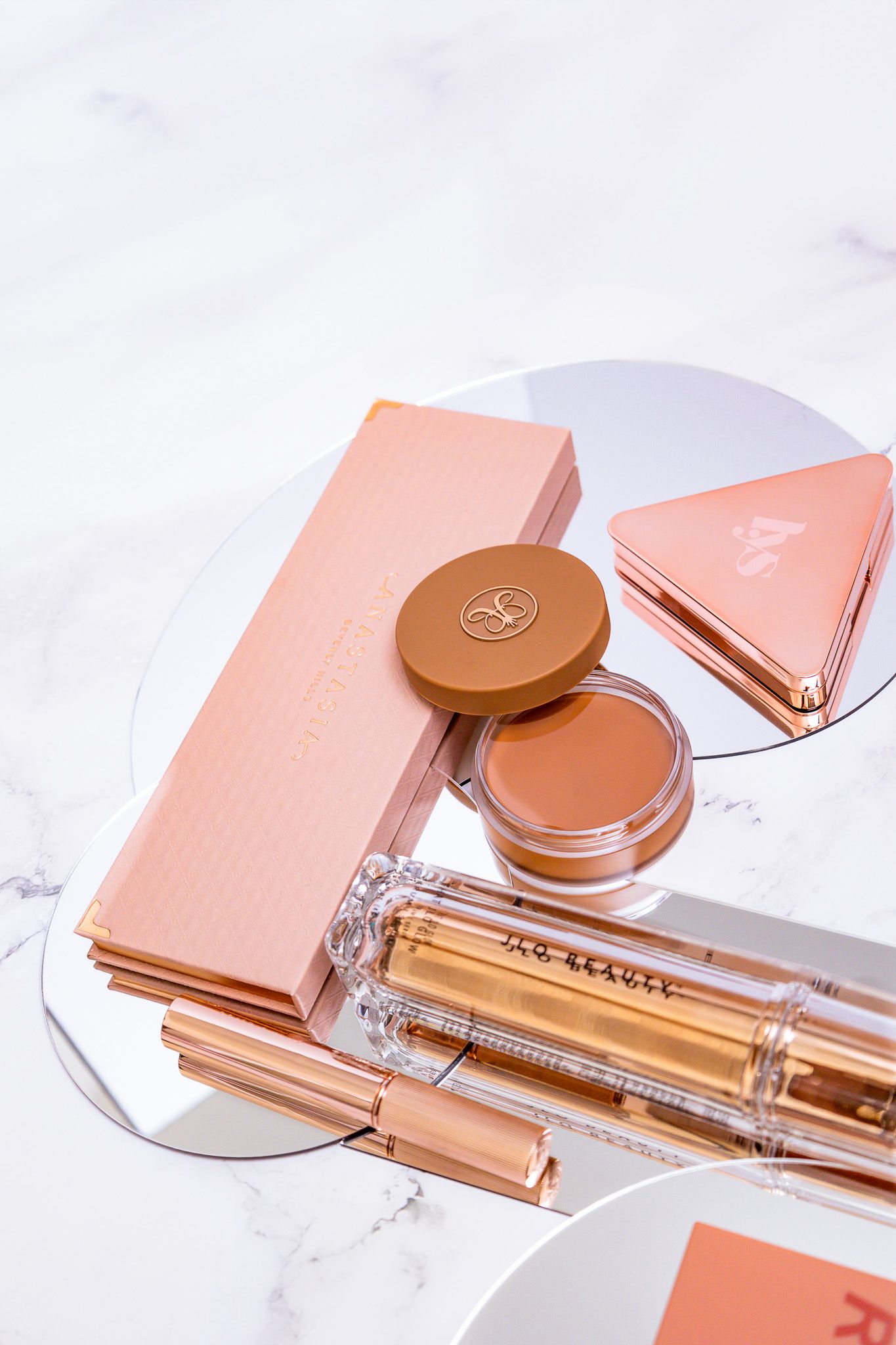 Rose gold beauty products and makeup accessories displayed on white marble background.
