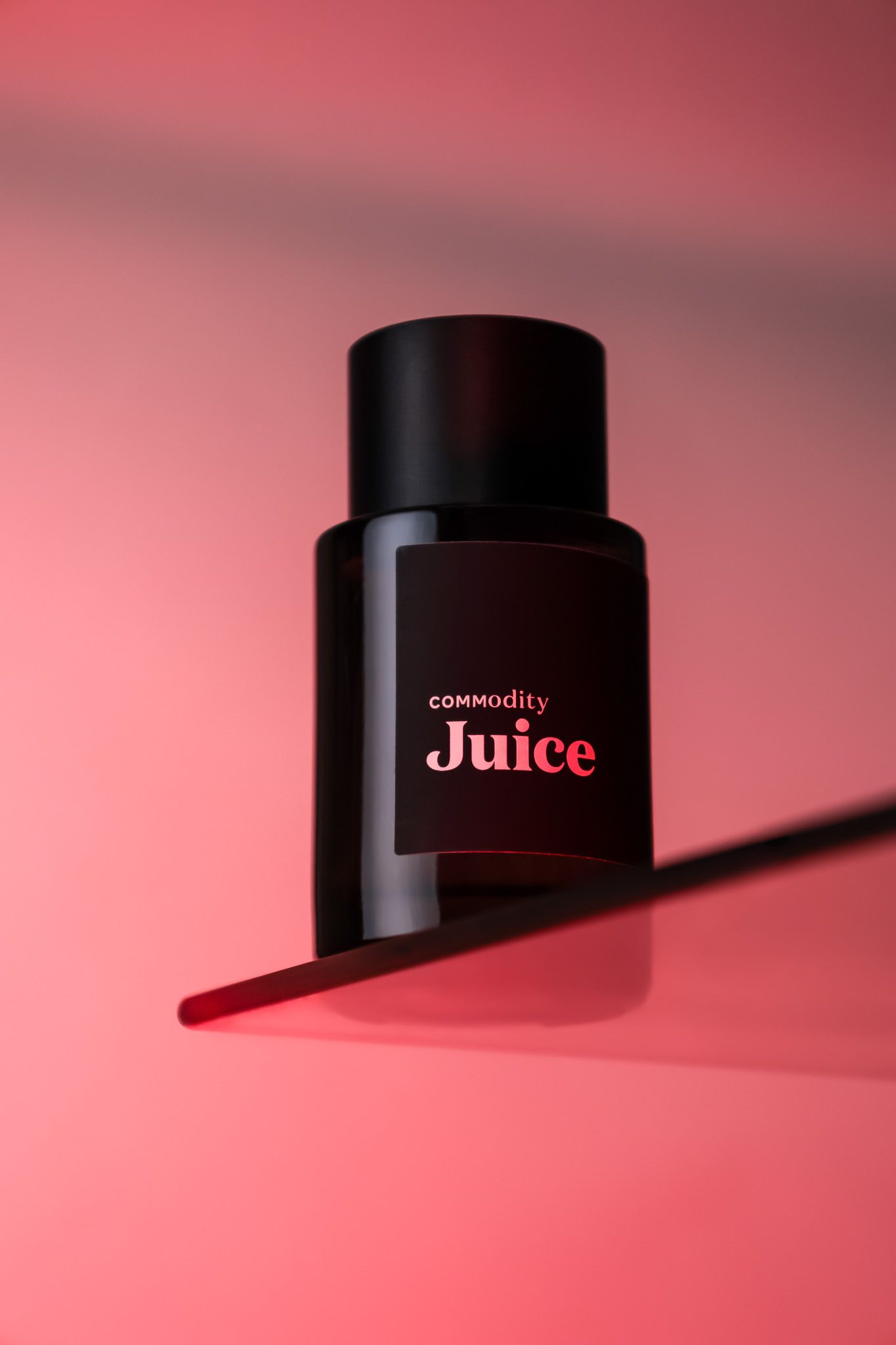 Black cylindrical bottle of Commodity Juice fragrance sits against a pink background with dramatic shadow.