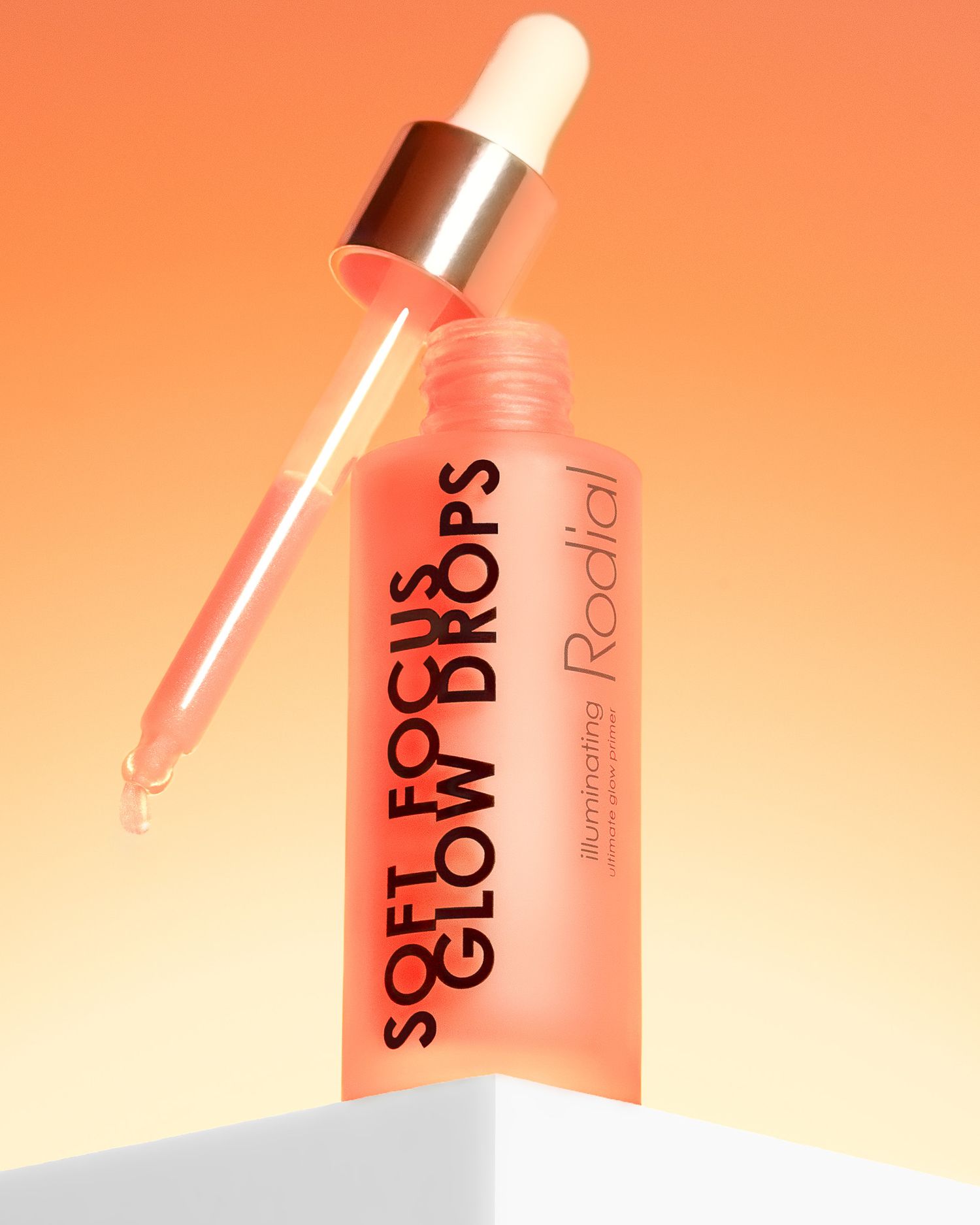 Rodial Beautys bottle of SOFT GLOW DROPS skincare serum with dropper against coral pink background showing liquid texture.