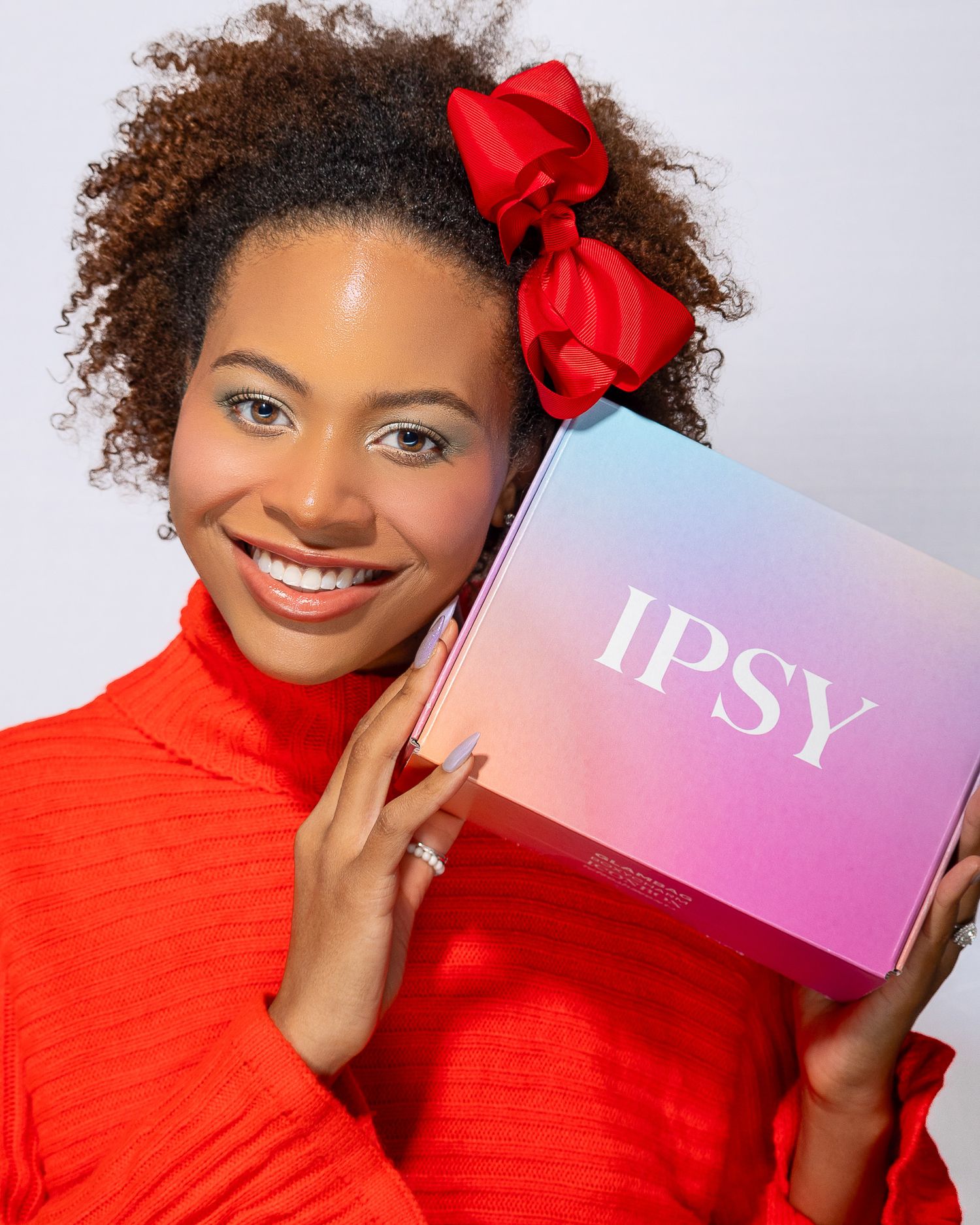 Beauty model in a bright red turtleneck sweater with curly hair and red bow accessories holds up an IPSY beauty subscription box.