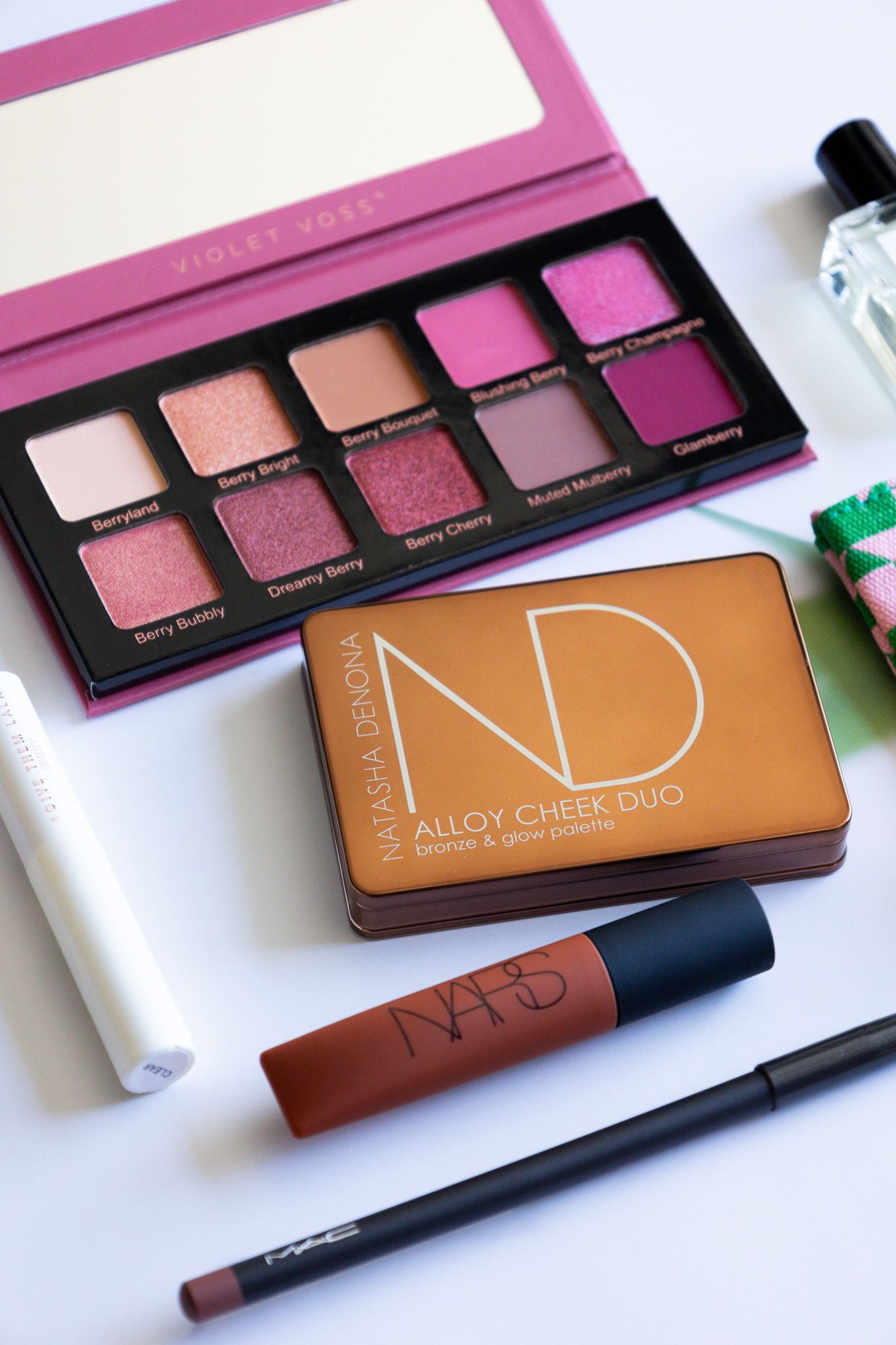 Flatlay of makeup products including NARS blush duo, pink eyeshadow palette, brown lipstick and black eyeliner on white surface.