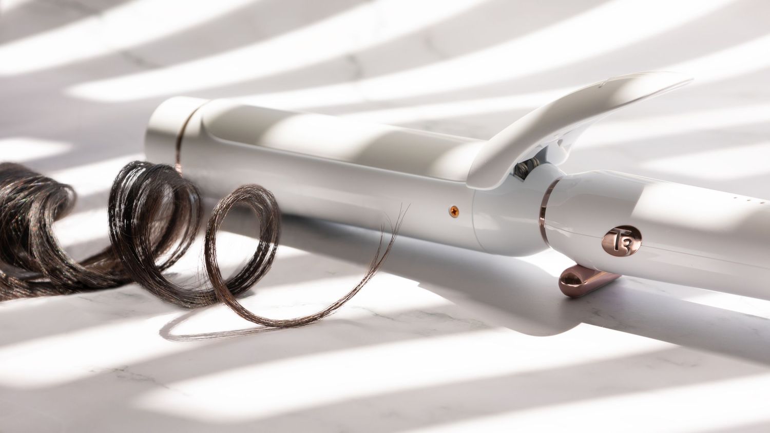 A silver curling iron with curved barrel casting shadows on a white surface.