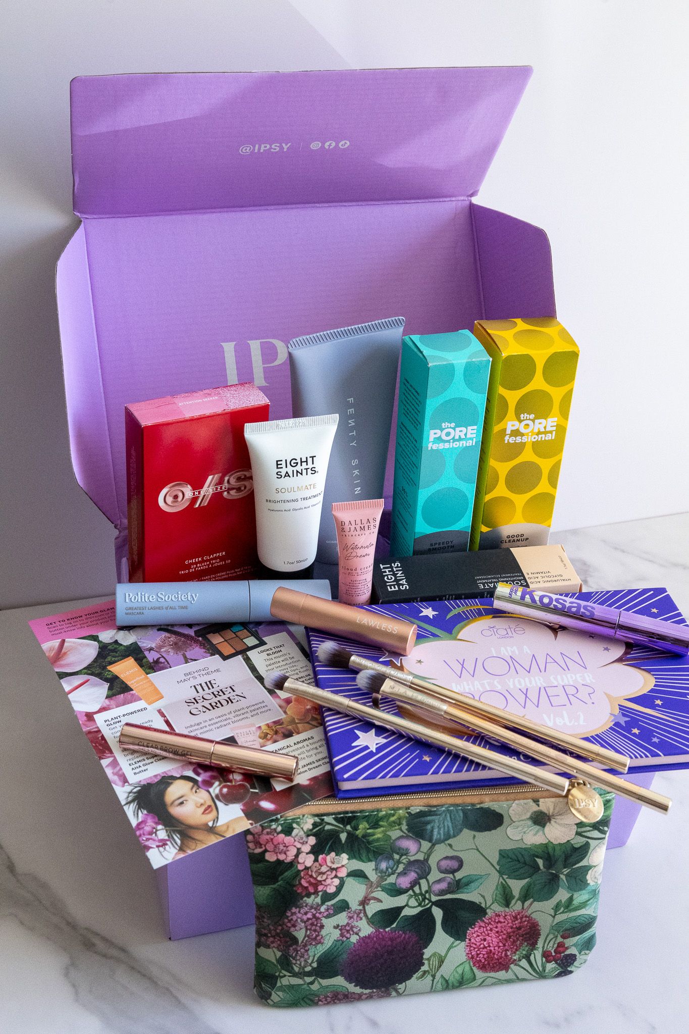 An open purple subscription box containing beauty products, magazines, and colorful packaging on a marble surface.