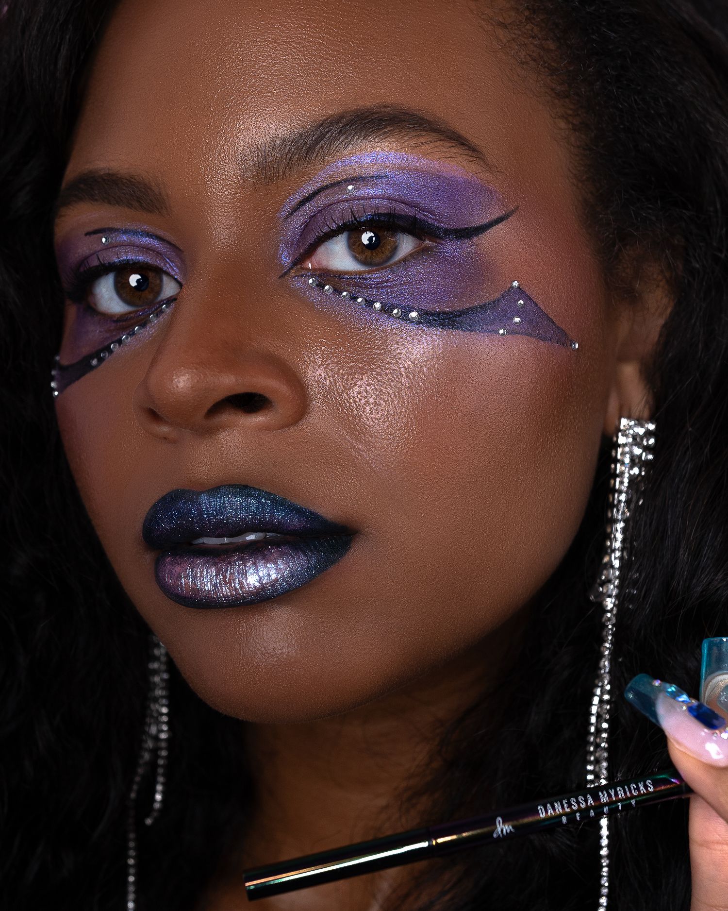 Close-up beauty shot showcasing dramatic purple and blue metallic makeup with rhinestone details.