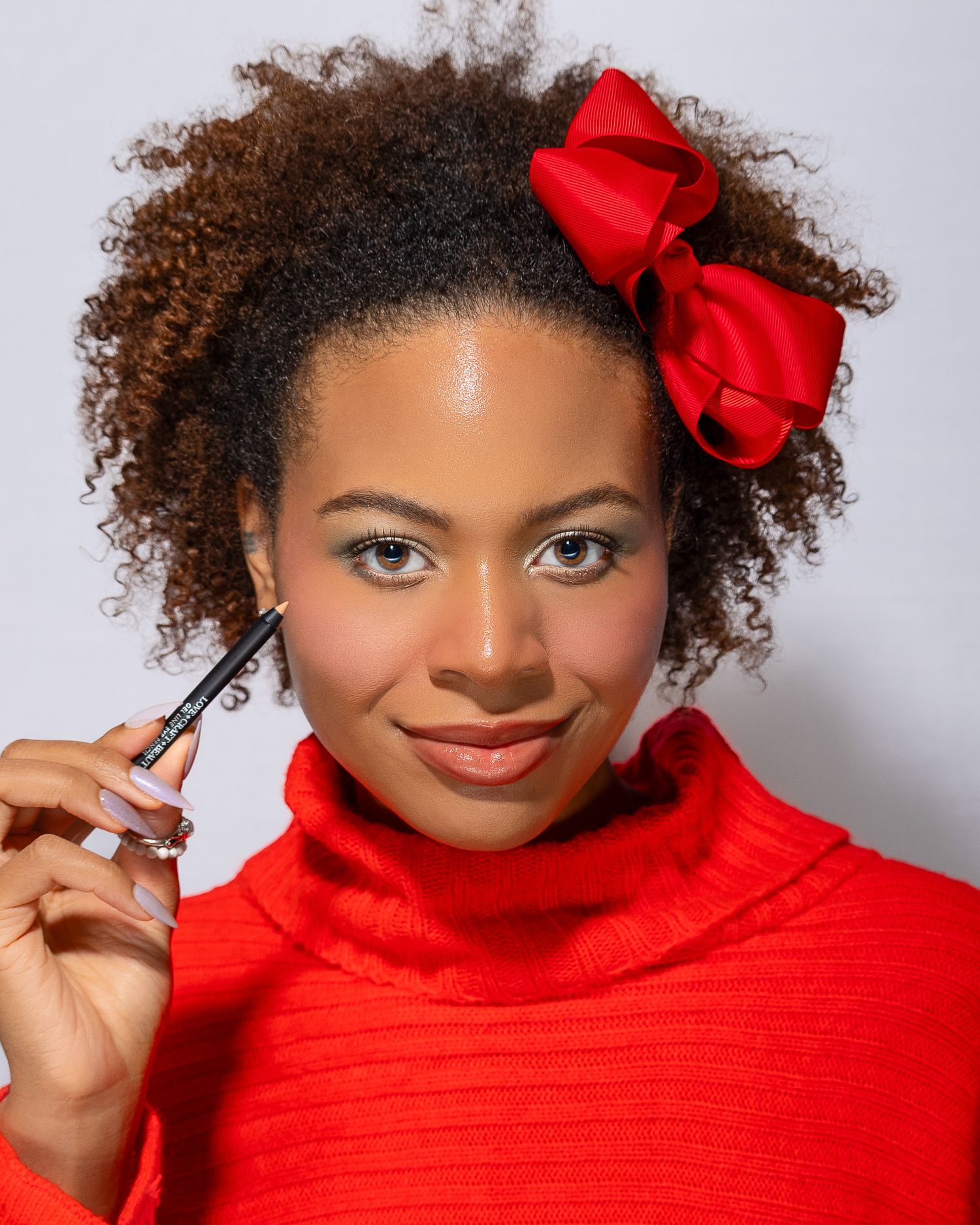 A beauty portrait showing natural curly hair styled with a red bow and red turtleneck sweater, holding a makeup product.