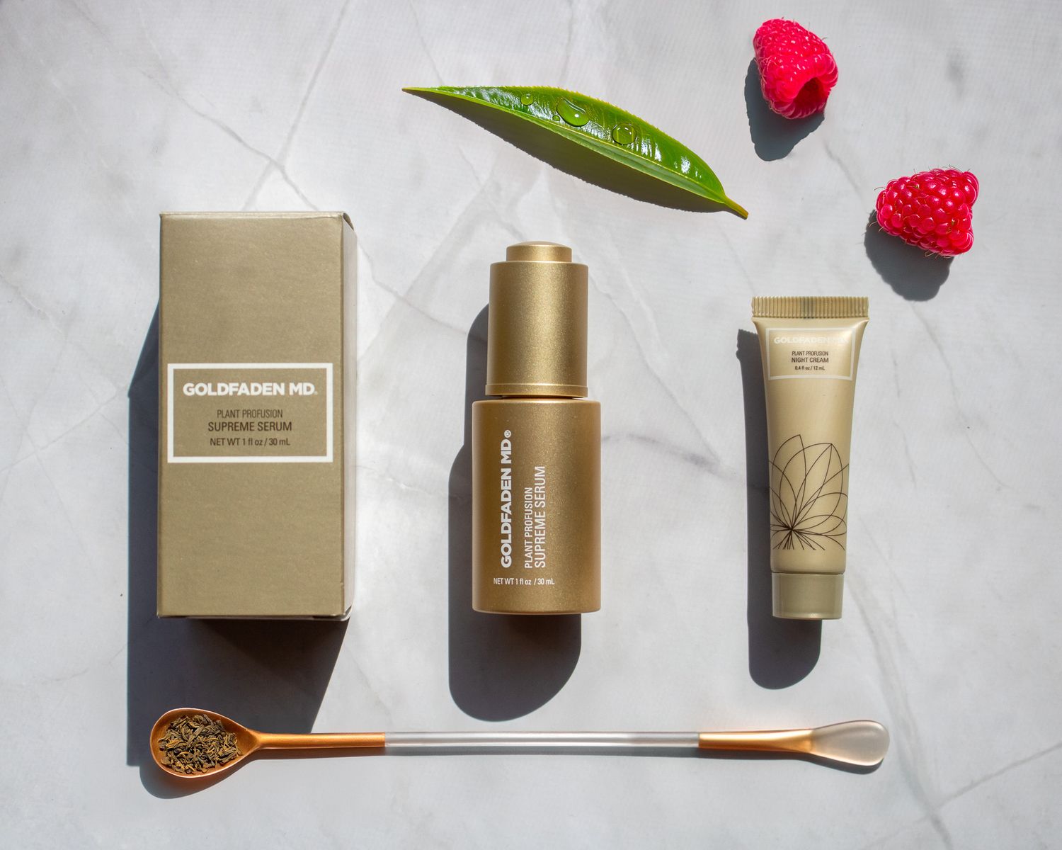 Beauty products arranged with gold packaging, pink flowers and green leaf on white marble surface.