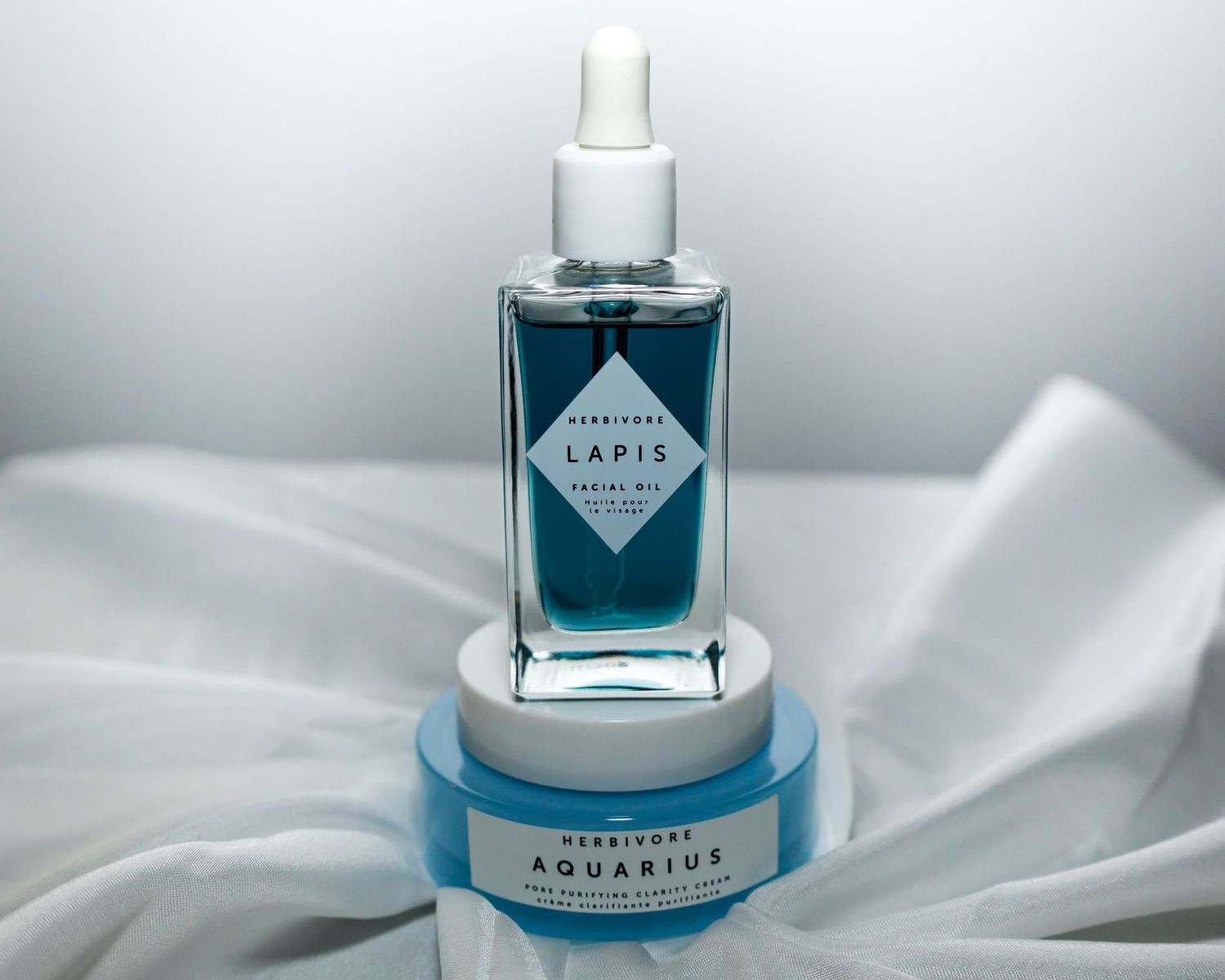 Blue glass bottle of Aquarius perfume displayed on white silk fabric with matching product jar.