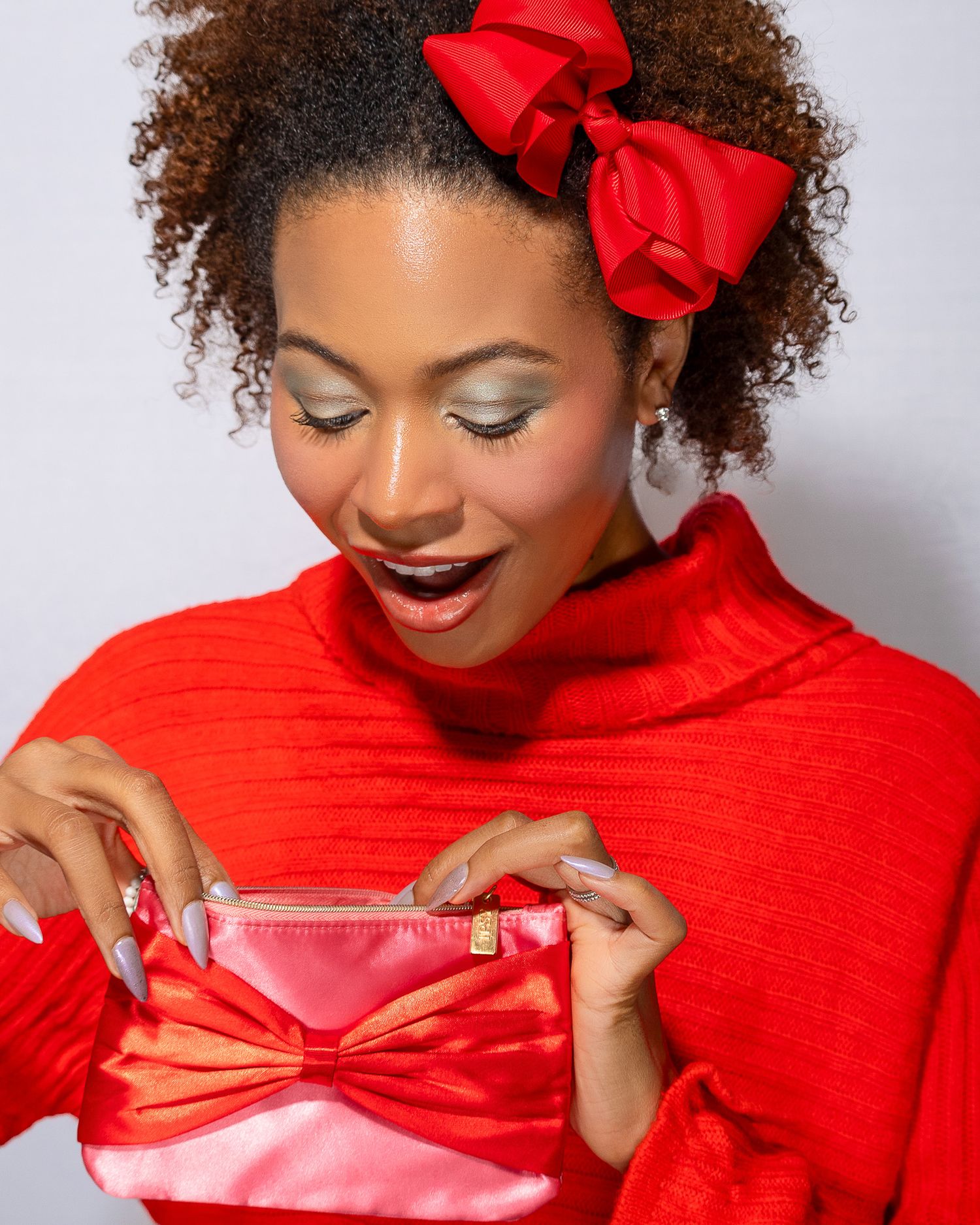 Woman wearing a red sweater and bow opens a pink gift box with an excited expression.