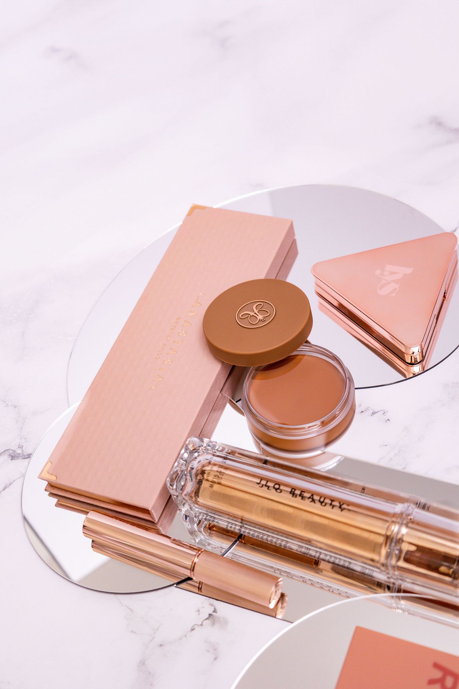 Feminine beauty products including makeup compact, lipstick and accessories in rose gold and neutral tones on marble surface.