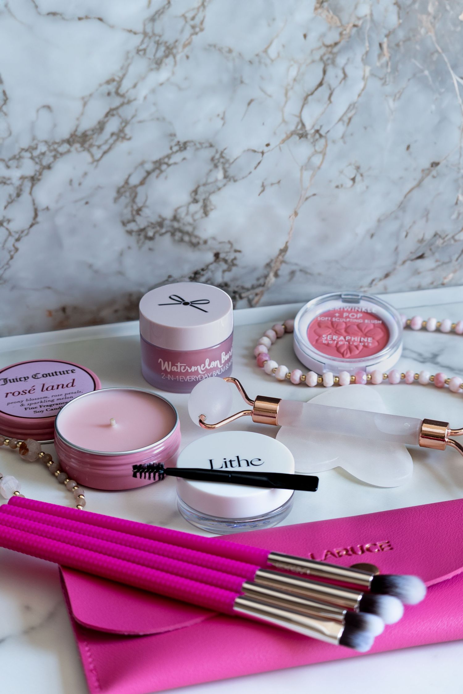 Pink makeup brushes and cosmetic products arranged on marble surface with pearls and rose gold accents.