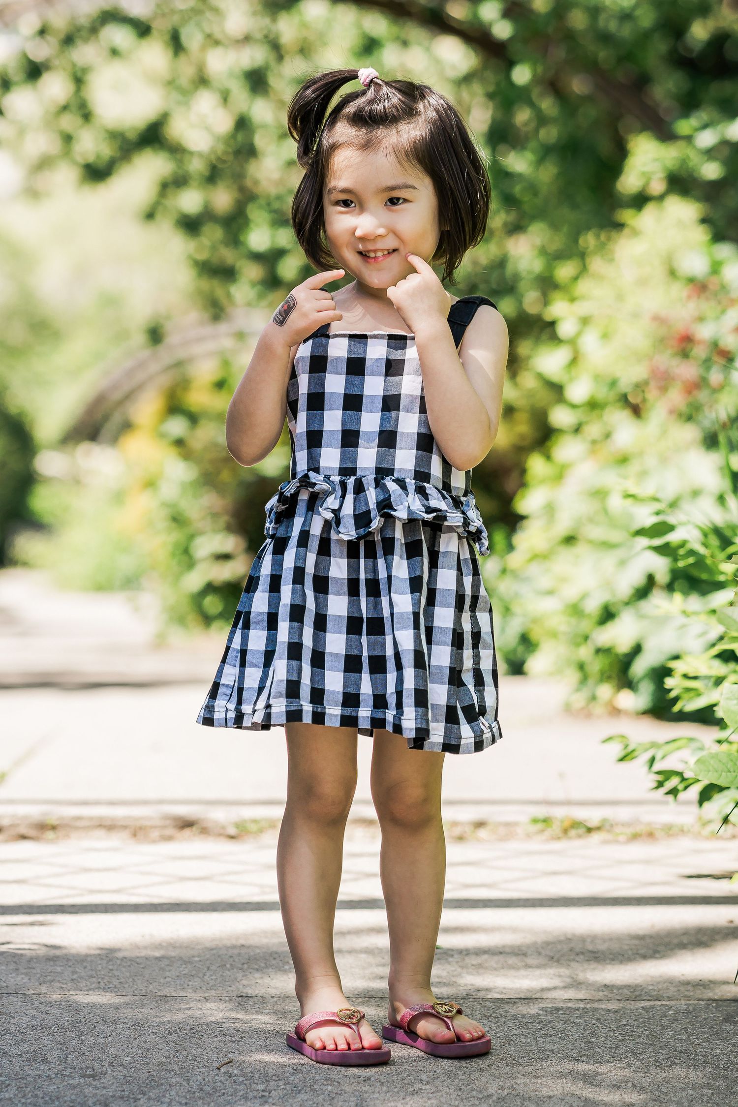 A young girl in a checkered sundress and pink flip flops strikes a pose in a leafy garden.