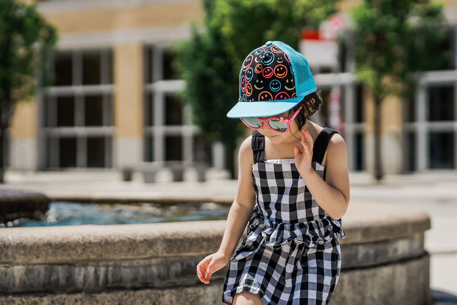 A young girl in a checkered sundress, a colourful blue hat, and pink sunglasses strikes a pose by a fountain.