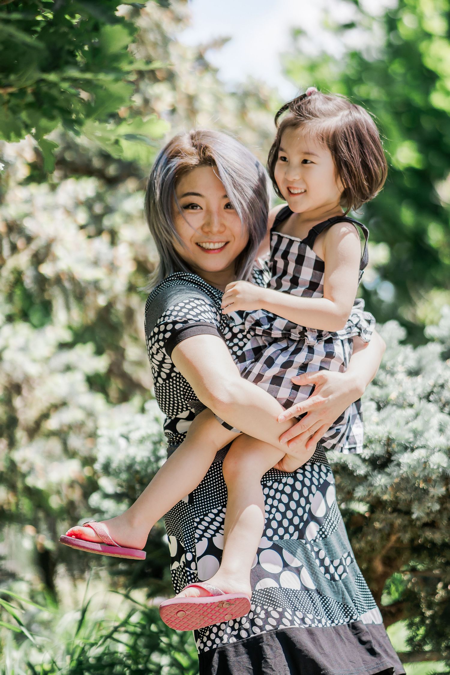 A woman with silver and blue hair holds up a young girl in a checkered sundress and pink flip flops as they stand in a leafy garden.