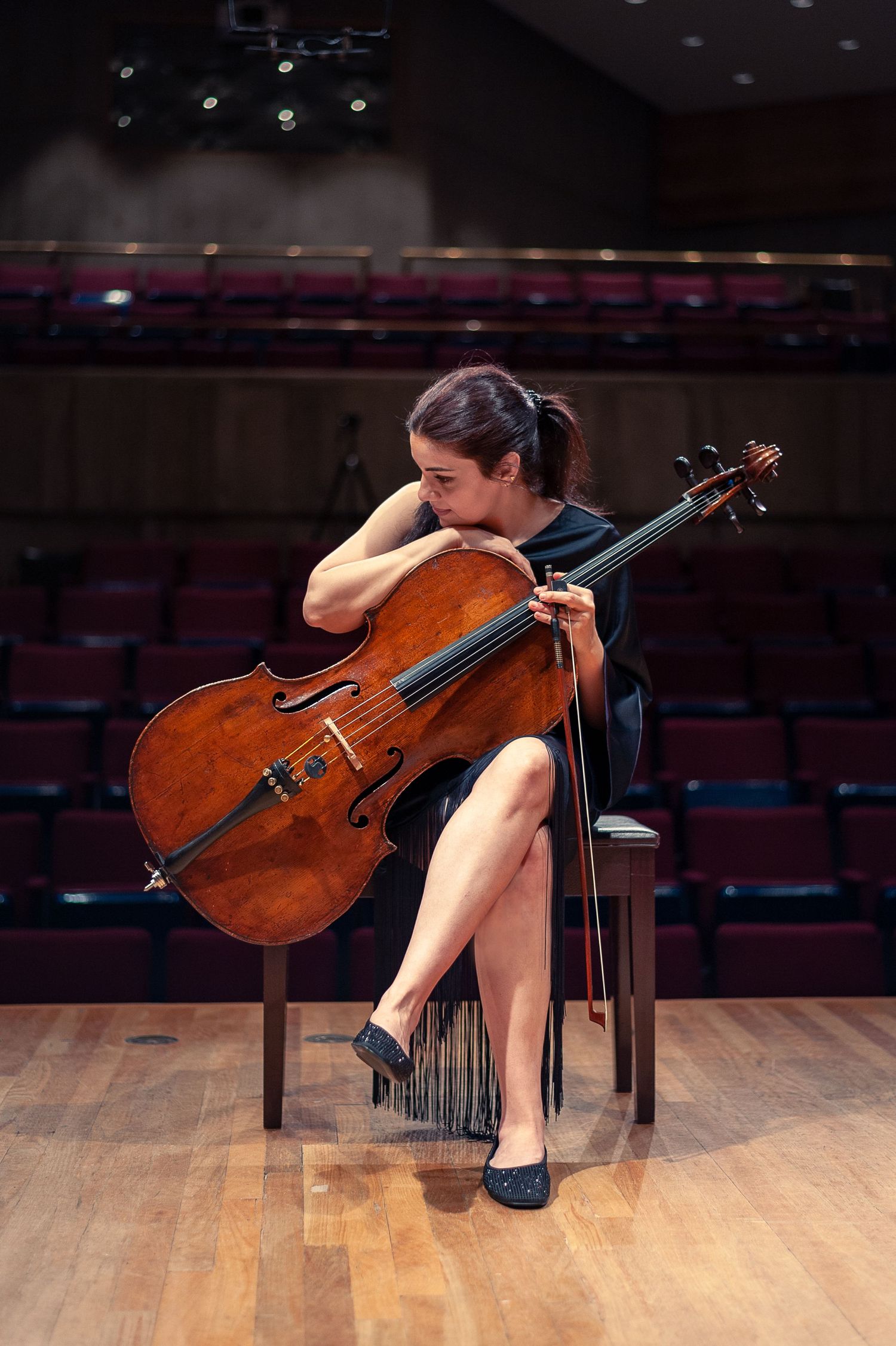 A woman with a serious expression sits in a black dress on stage holds her cello sideways.