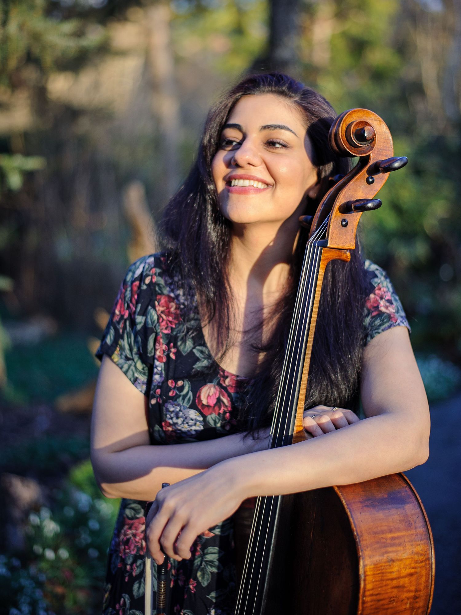 A woman smiles in a sunny garden holding a cello.