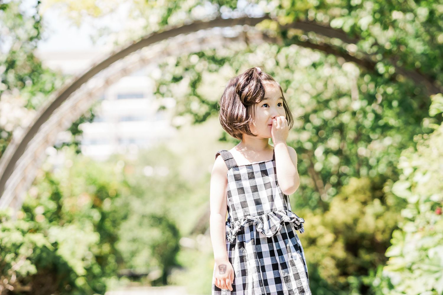 A young girl in a checkered sundress thoughtfully looks off to the right in a leafy garden, framed by an arch.