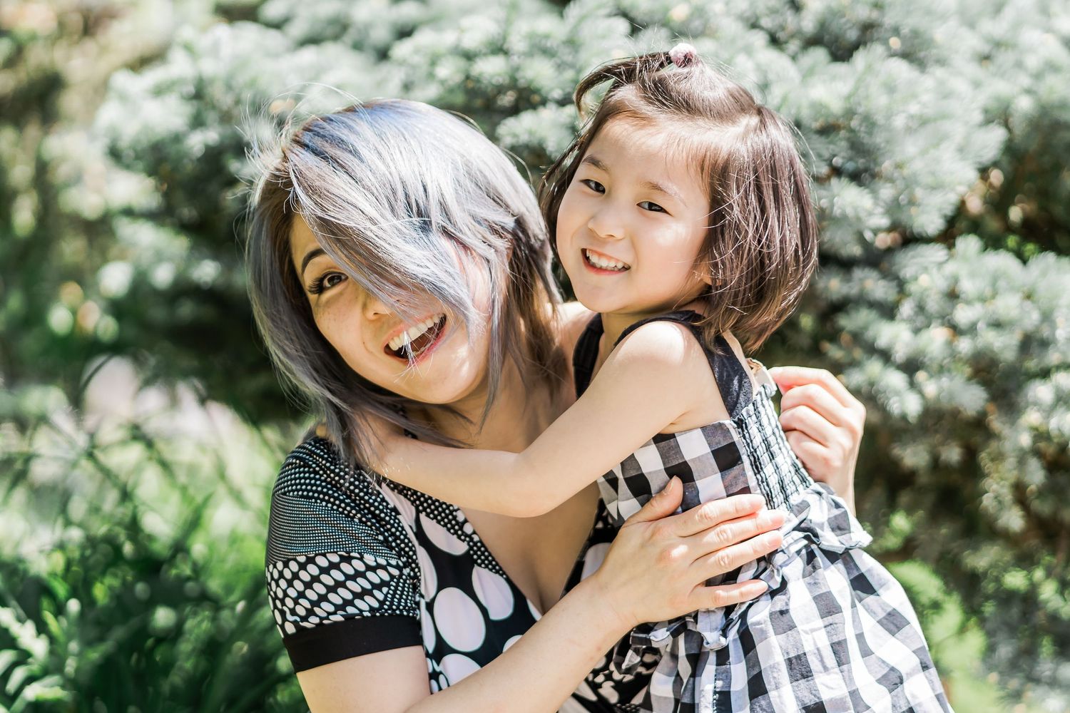A woman with silver and blue hair hugs a young girl in a checkered sundress as they sit in a leafy garden.