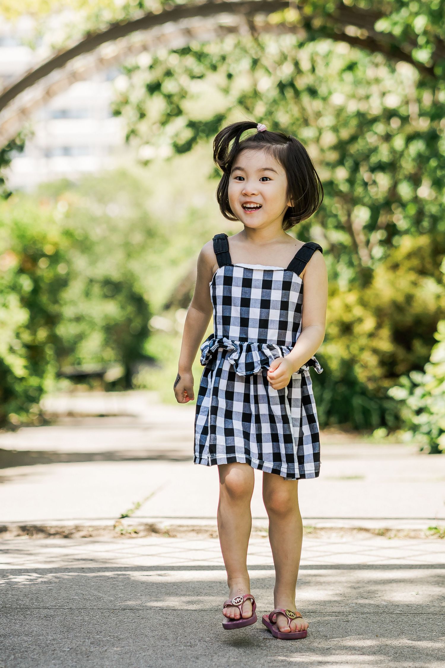 A young girl in a checkered sundress smiles and walks through a leafy garden.