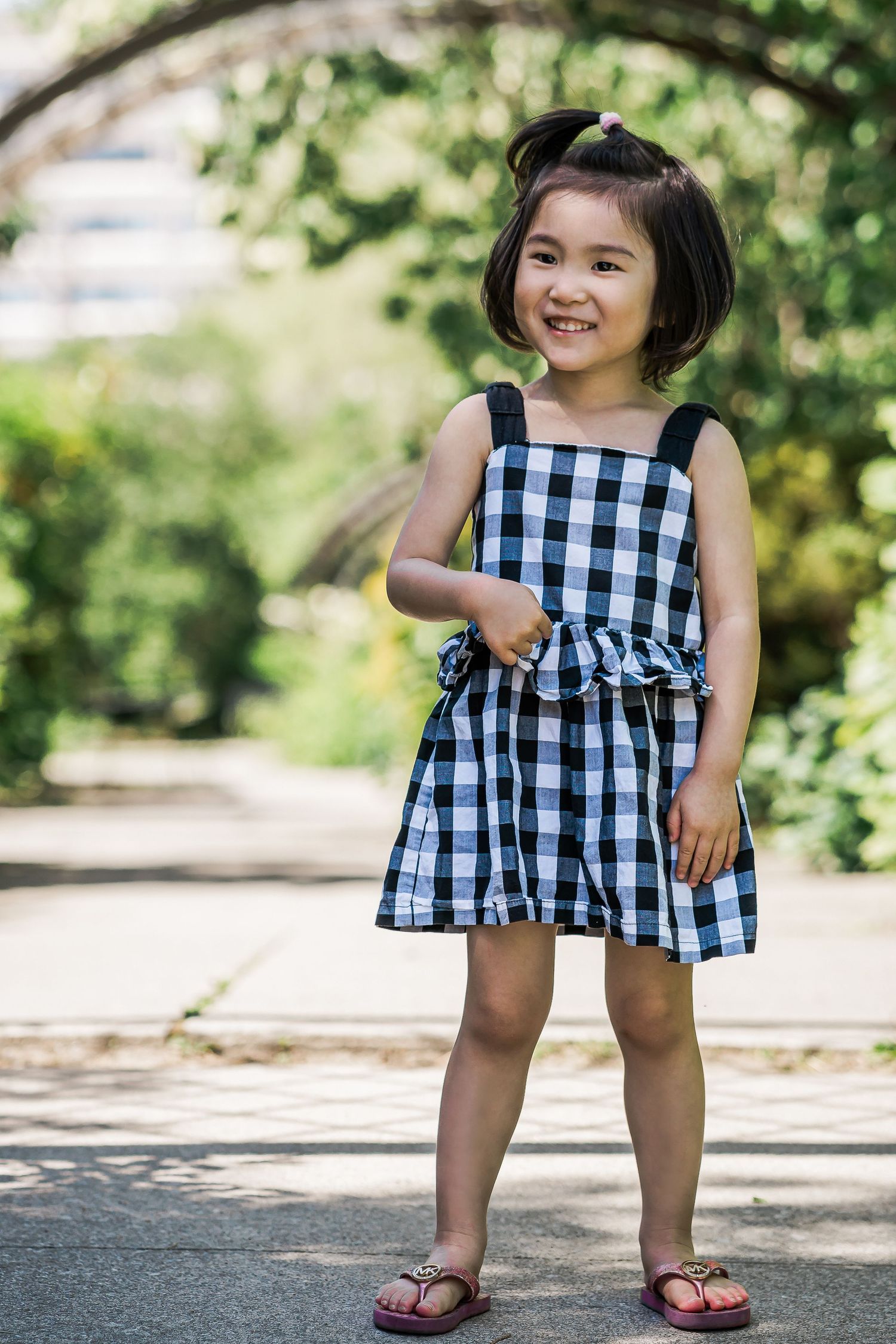A young girl in a checkered sundress points to her belly and smiles in a leafy garden.