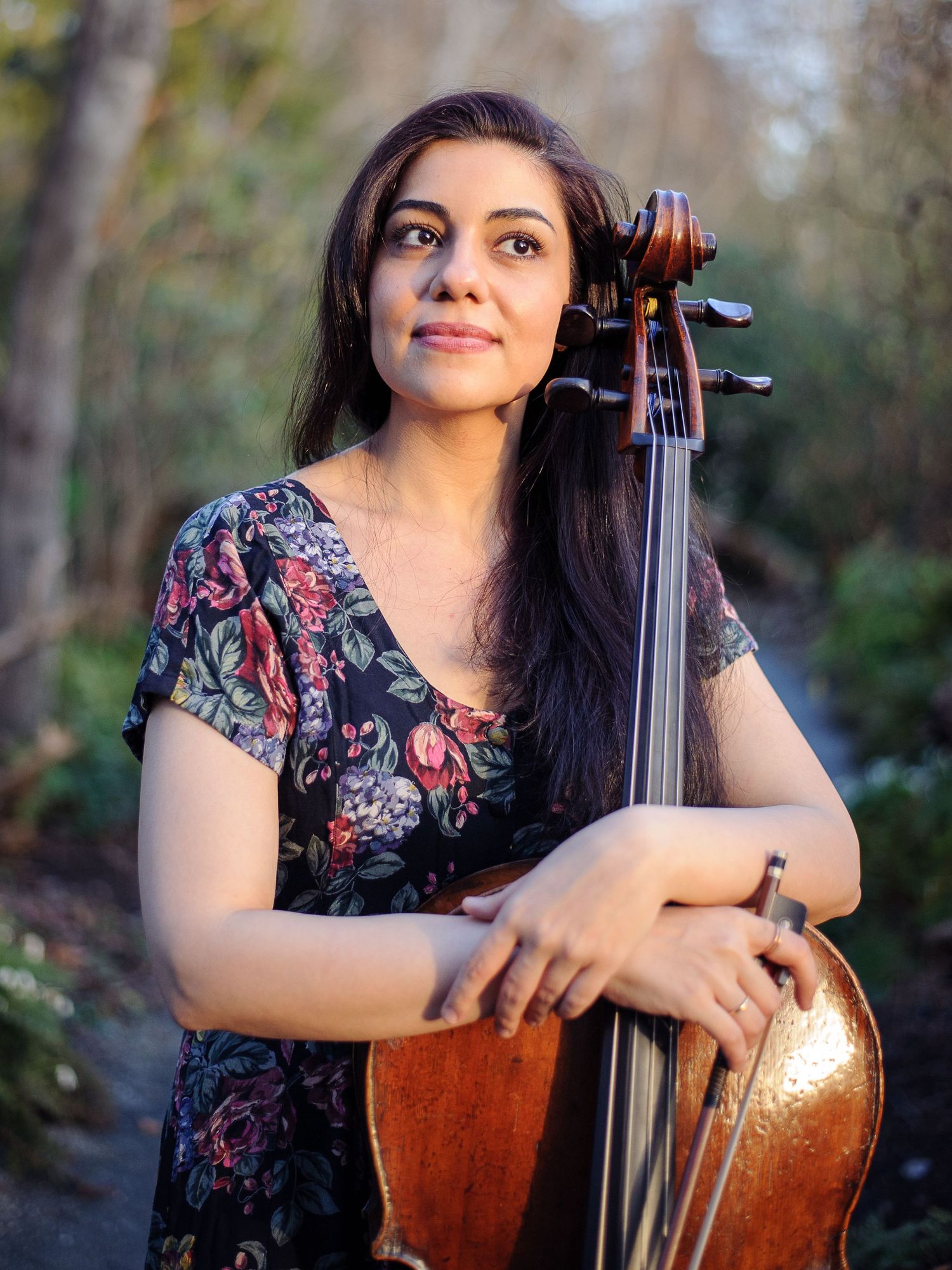 A woman in a flowery dress hugs her cello in a sunny garden.